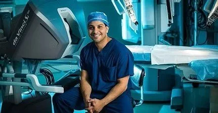 Smiling healthcare professional in navy scrubs sitting in a hospital or surgical room with advanced medical equipment.