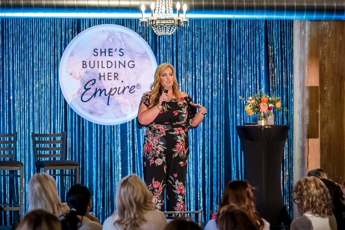 A woman in floral jumpsuit speaking into a microphone on stage with a large screen behind her that reads "She's Building Her Empire". The stage has blue metallic curtains, a chandelier overhead, and a vase of flowers on a tall stand.