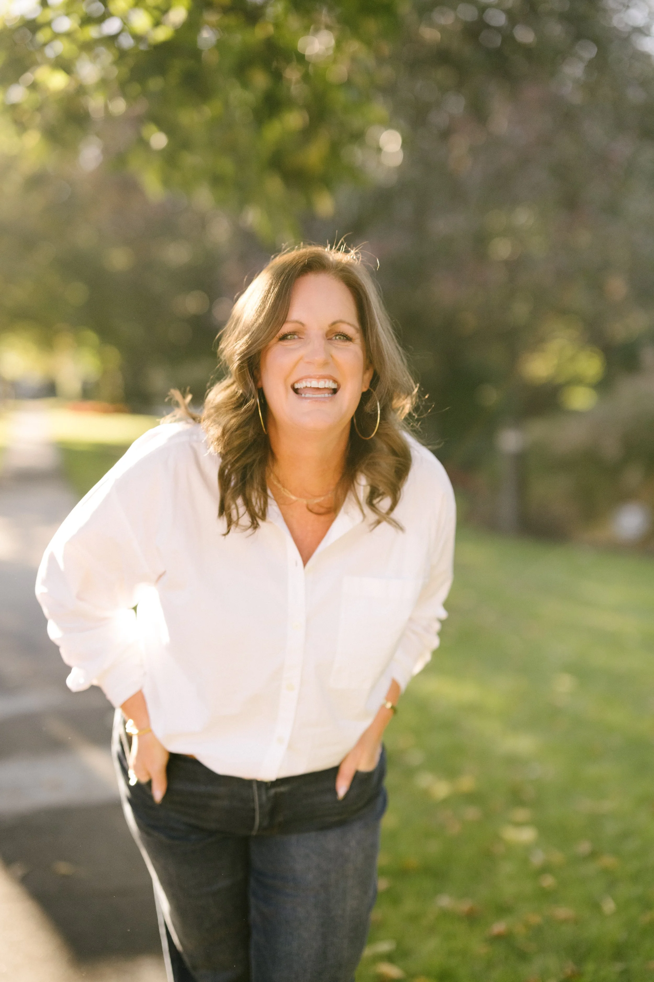 A woman smiling and leaning forward outdoors, wearing a white shirt and hoop earrings, with blurred trees and a pathway in the background.