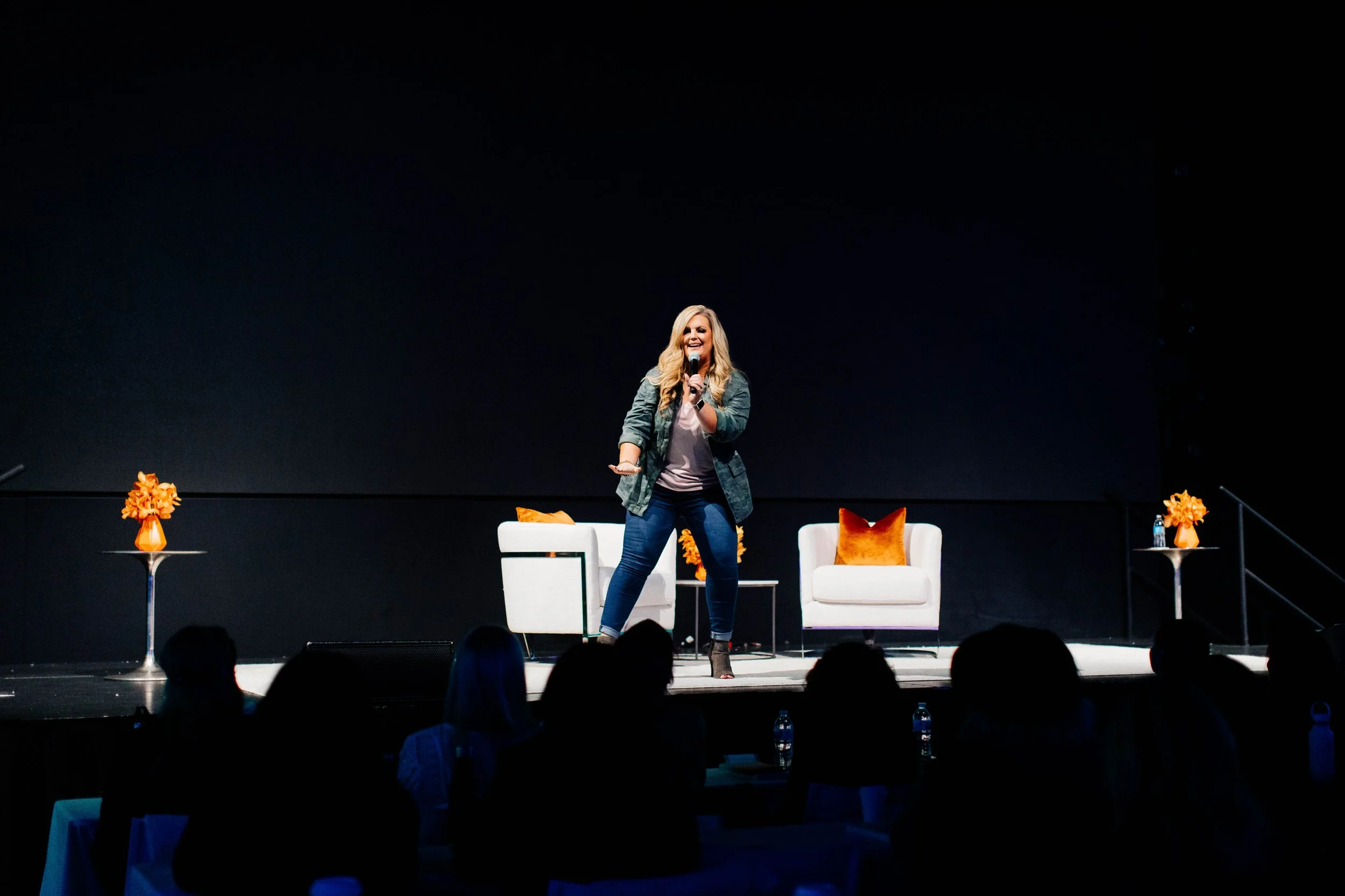 A woman with long blonde hair holding a microphone, smiling, standing on a stage with a dark background. there are two white chairs with orange pillows and small tables with orange flowers on stage, and the audience is visible in the foreground.