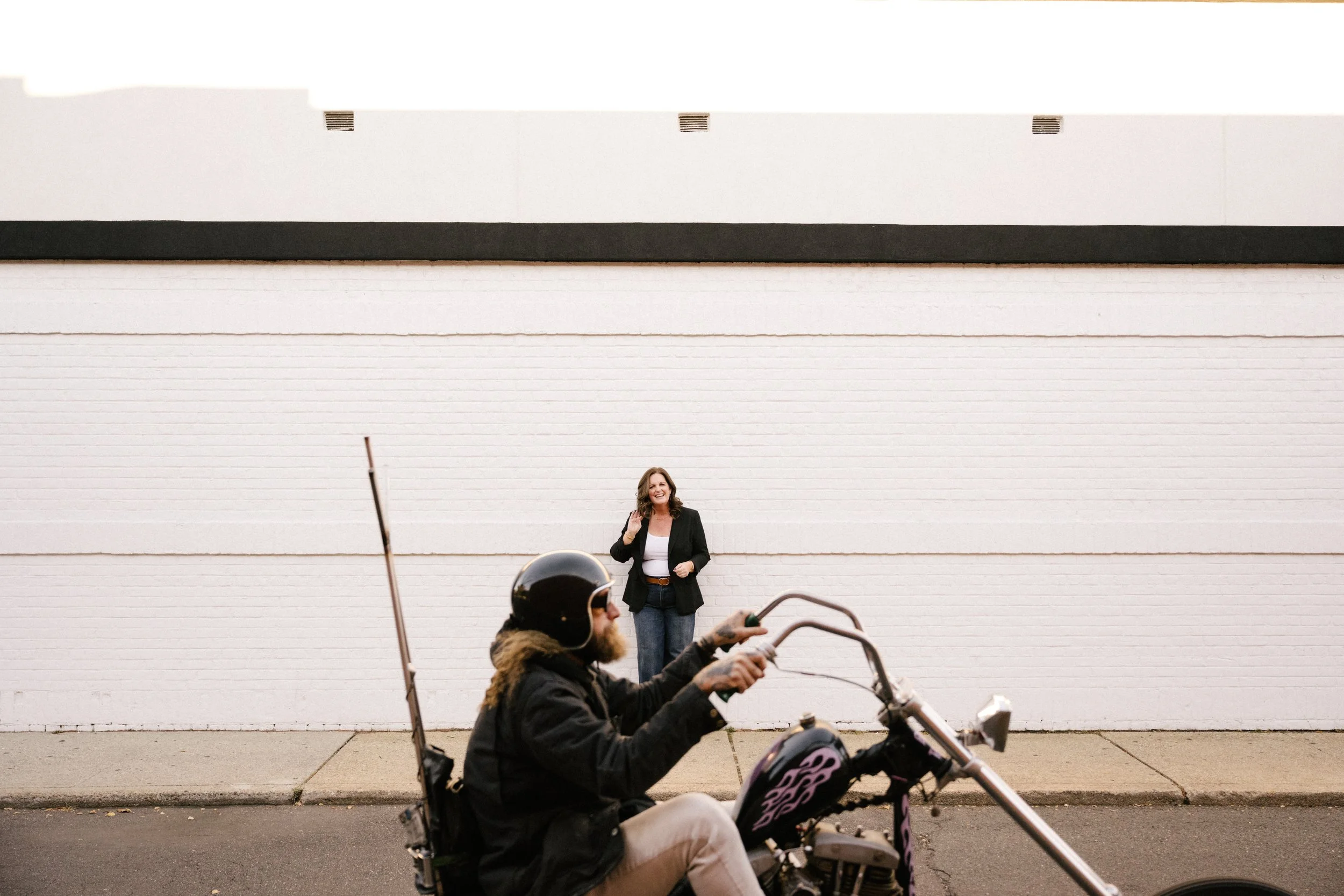A woman standing and smiling against a white brick wall, with two people riding motorcycles in the foreground.