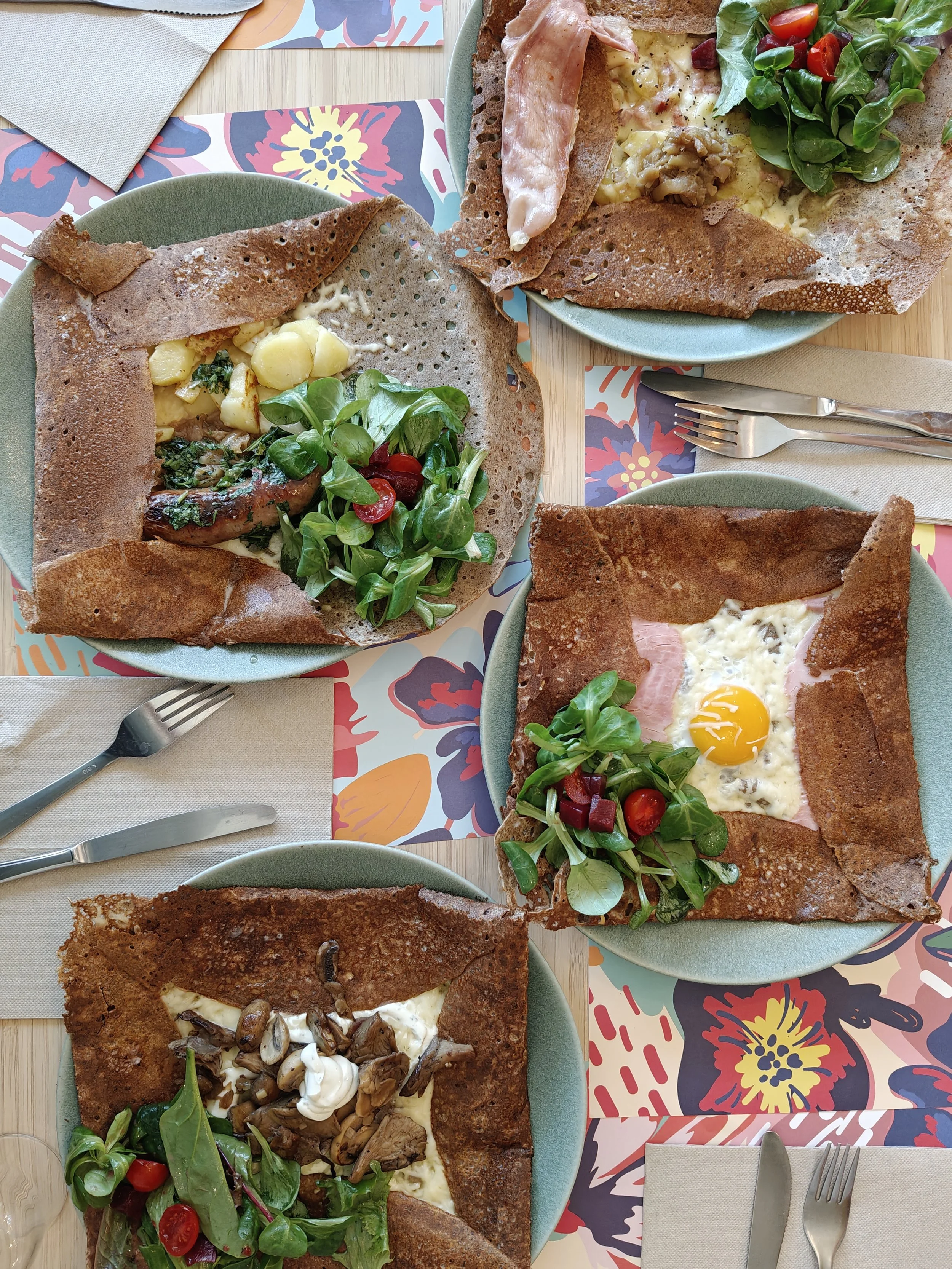 Quatre galettes bretonnes de sarrasin avec différentes garnitures, comprenant un œuf au plat, du jambon, du fromage, des champignons, de la salade verte, des tomates cerises, et des petits légumes sur un fond de napperons en papier à motifs floraux.