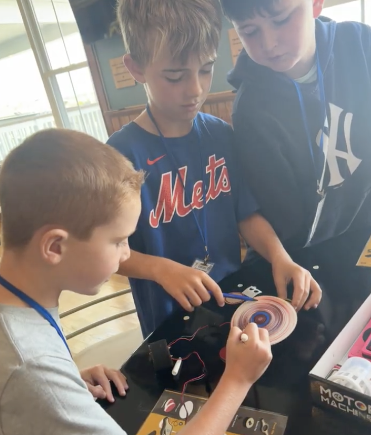 Three boys working on a science or engineering project involving a spinning disc and electrical wires at a table.