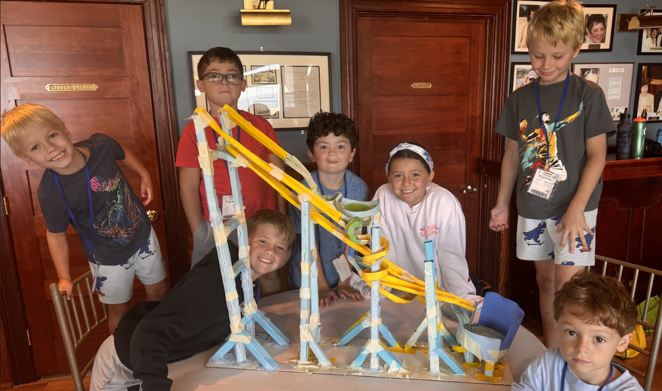 Group of children gathered around a table with a colorful roller coaster model made from foam and plastic tracks. The children are smiling and appear proud of their project inside a room with wooden walls and framed photographs.