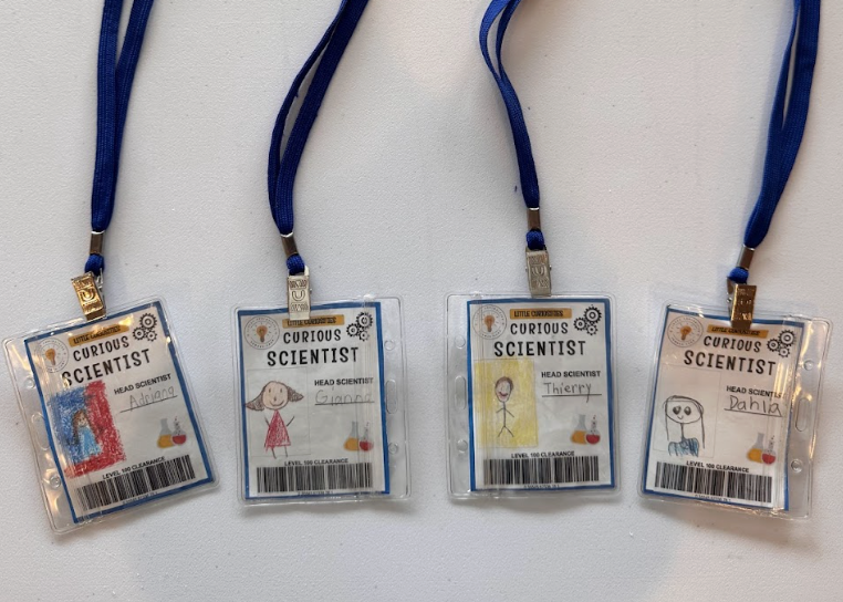 Four toy science badges with colorful drawings and names, hanging on blue lanyards.