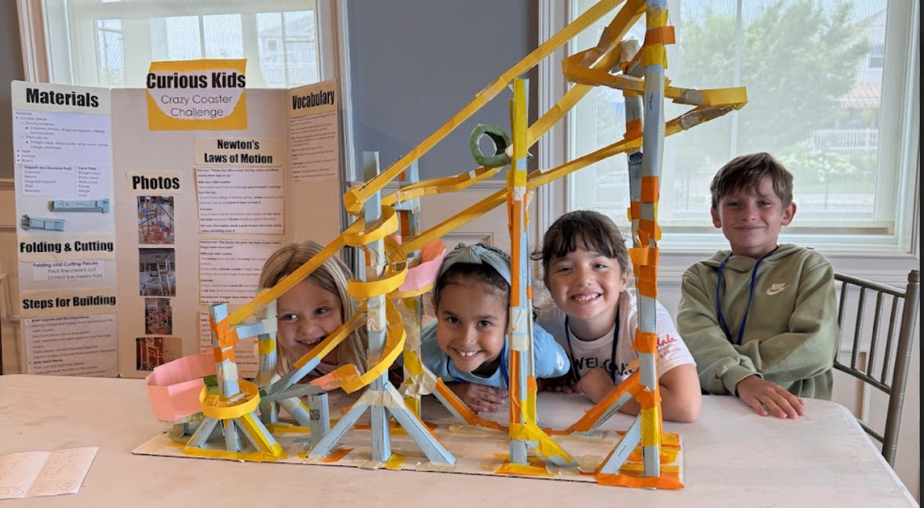Four children are seen smiling behind a large model of a roller coaster made from paper or cardboard, with a background display board on the left and windows behind them.