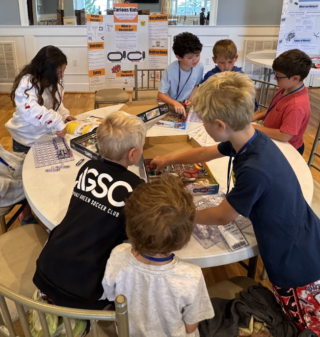 Children gathered around a table participating in a STEM activity or science experiment, with informational posters in the background about electricity and inventions.