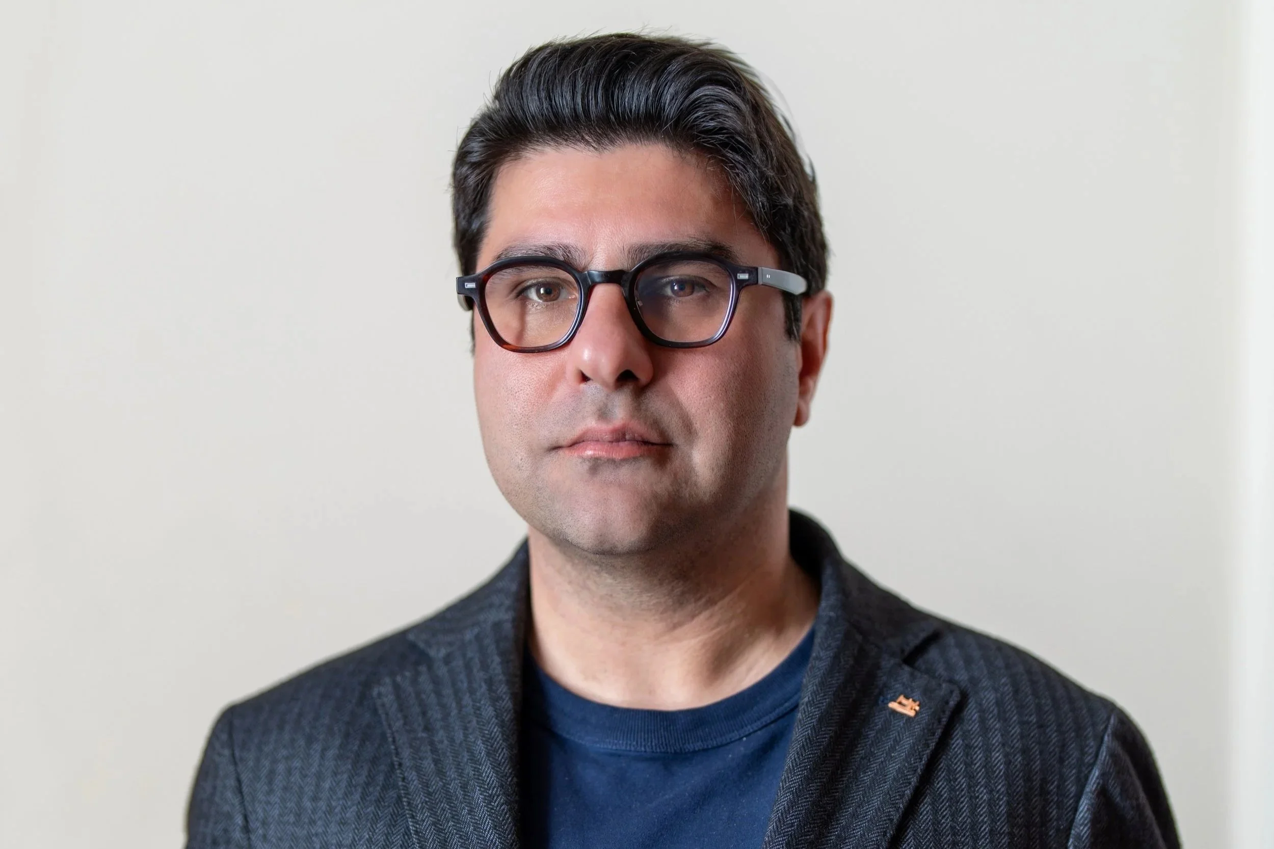 Portrait of a man with dark hair, glasses, wearing a dark blazer and a navy shirt, standing against a plain light-colored background.