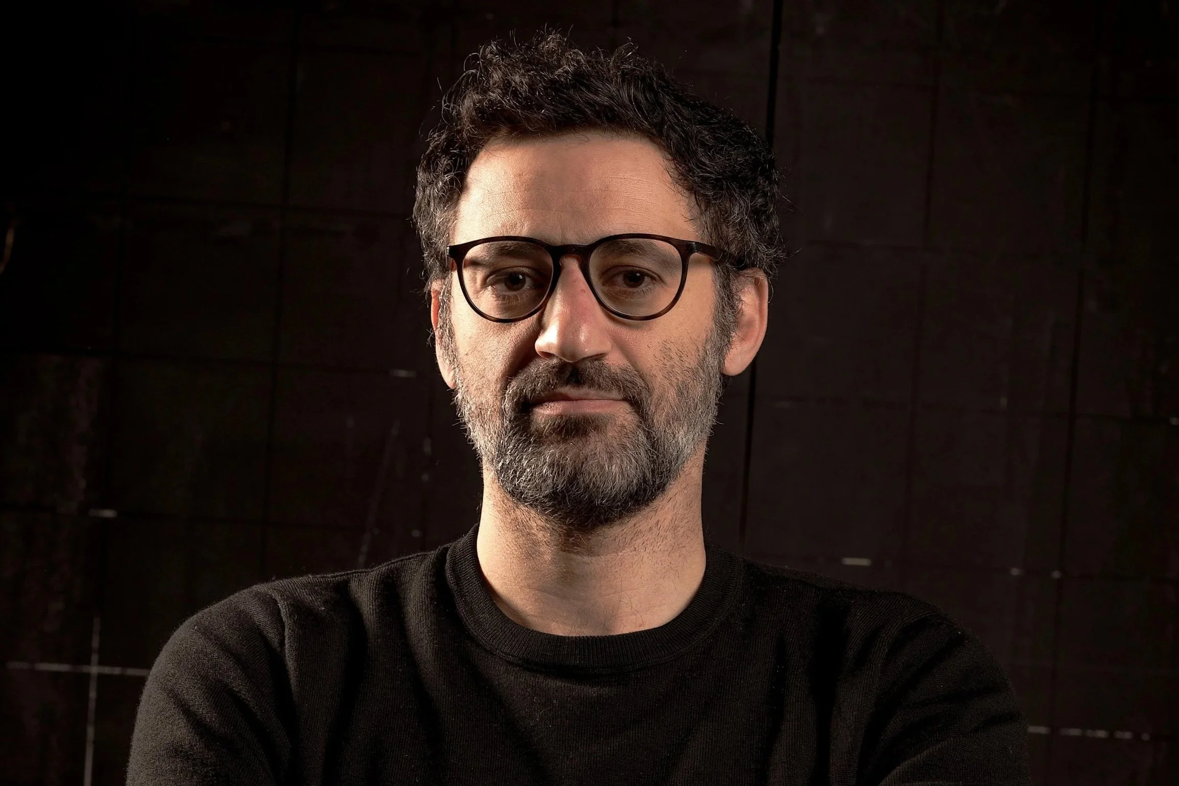 A man with curly dark hair, beard, and glasses wearing a black shirt, standing against a dark background.