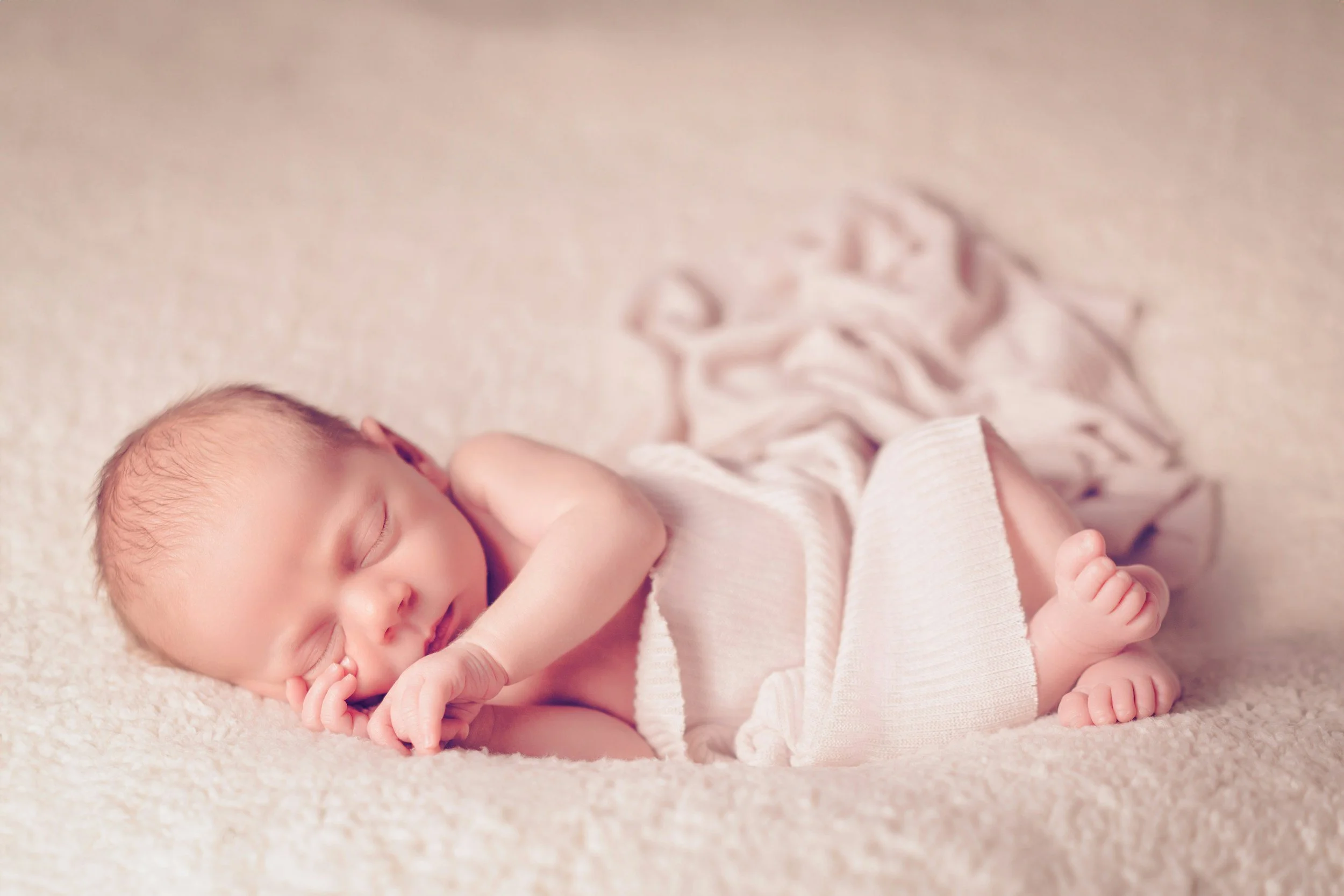 A sleeping baby laying on a soft, cream-colored blanket with hands near face and curled up with knees to chest.