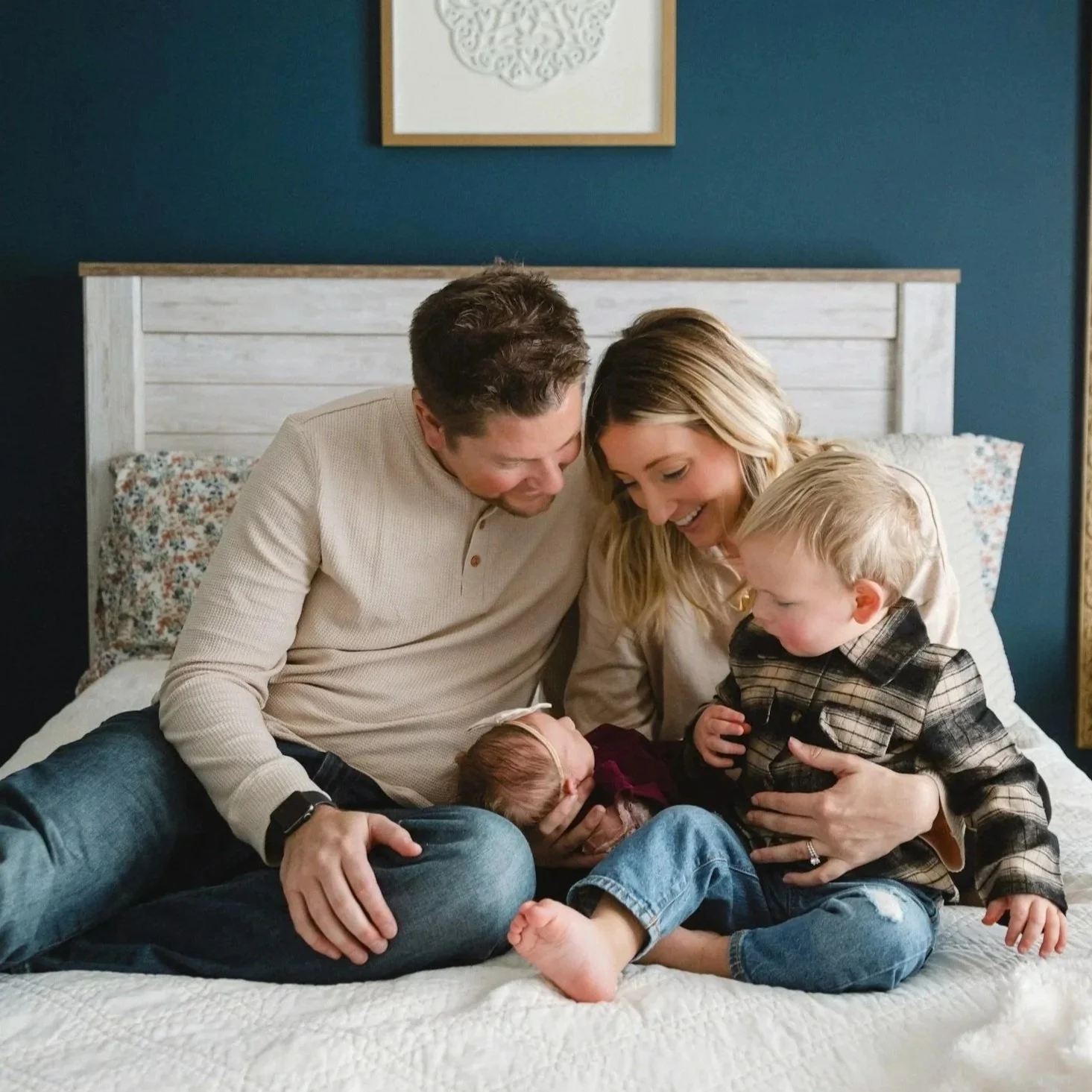 A family of five, including two young children and two adults, sitting on a bed and looking at an infant with a bandage on its head, showing affection and joy in a cozy bedroom setting.