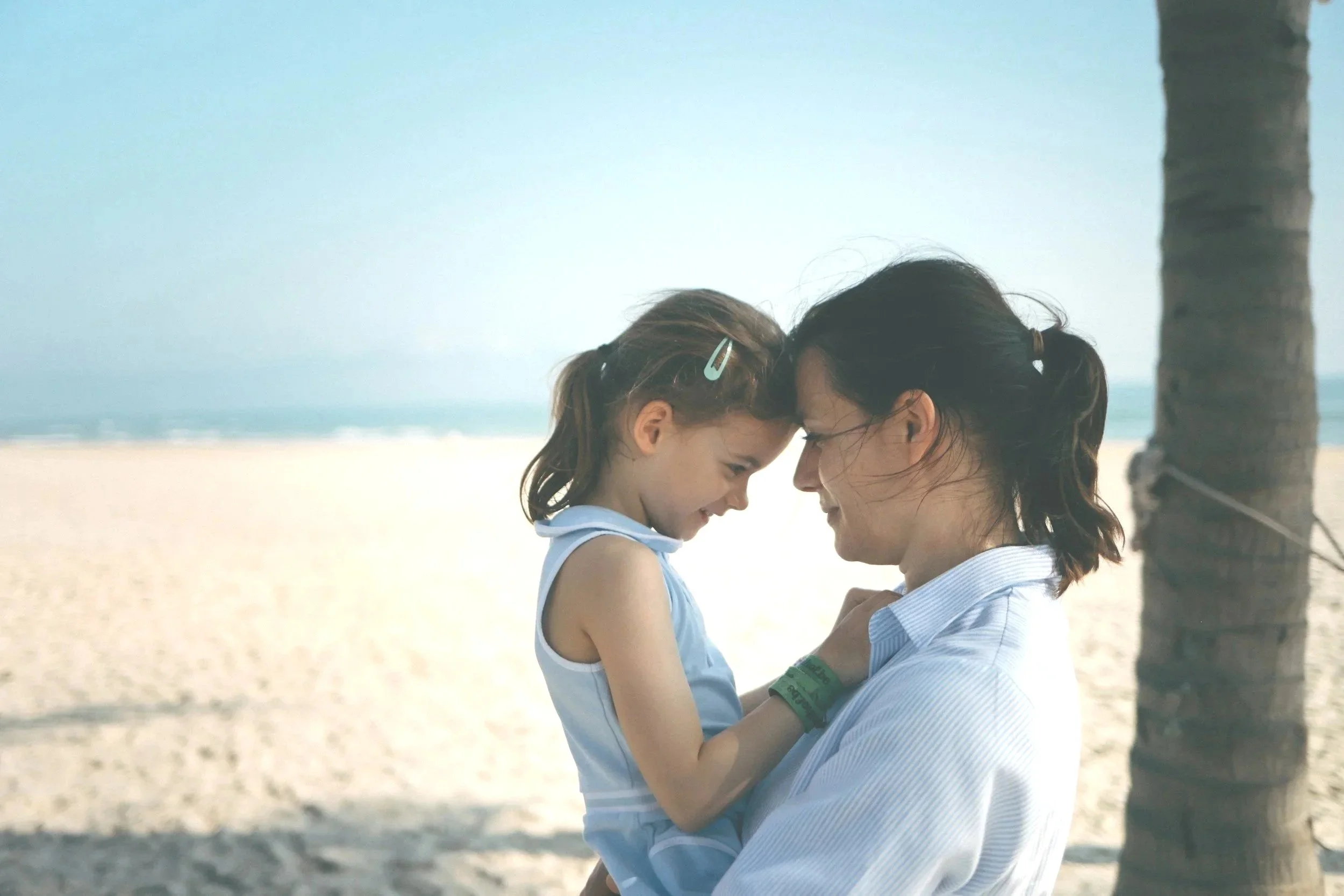 A father and daughter sharing a tender moment on the beach with an ocean and palm tree in the background.