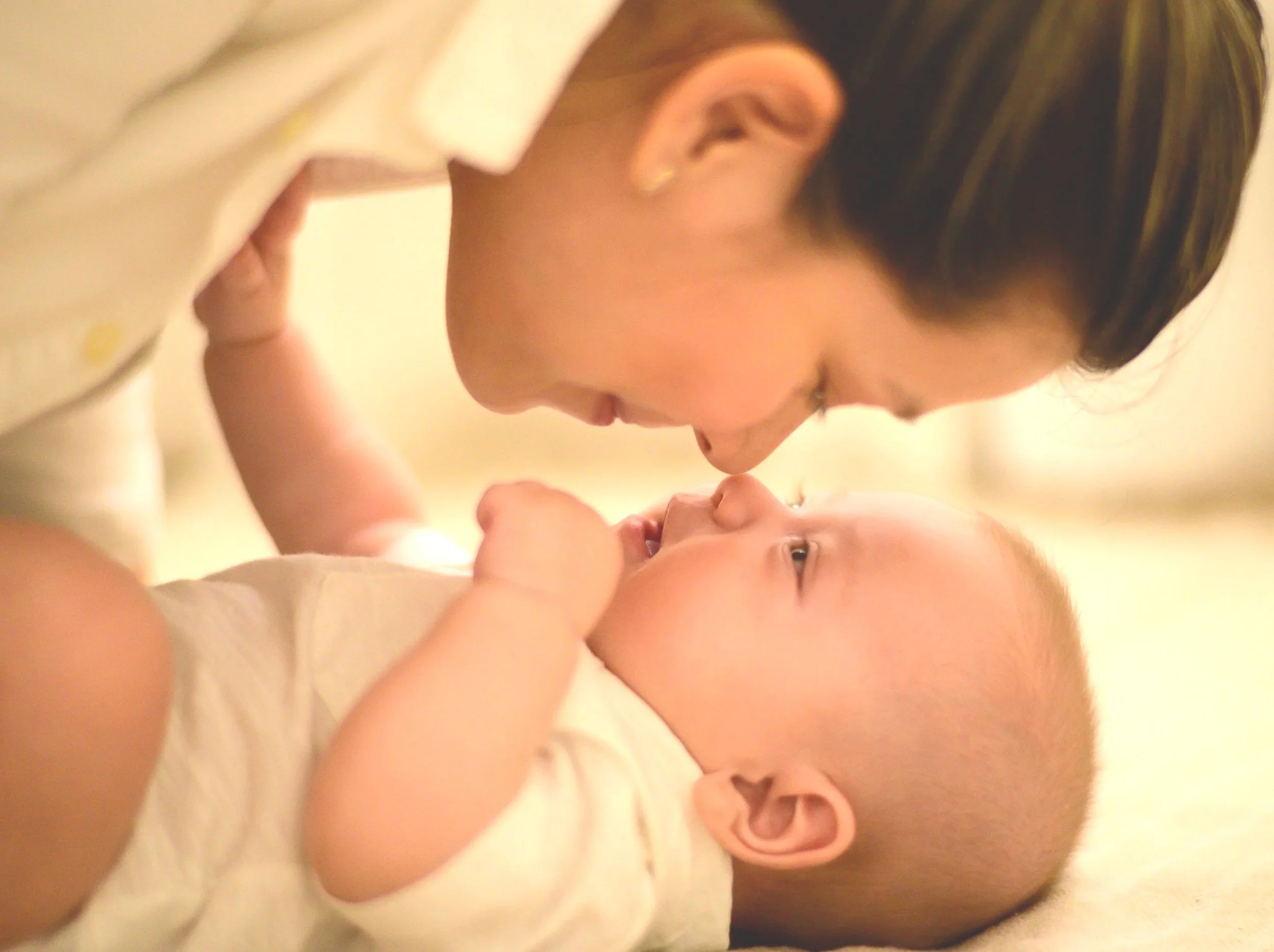 A woman and a baby are lying down face to face, smiling and touching noses in a warm, intimate moment.