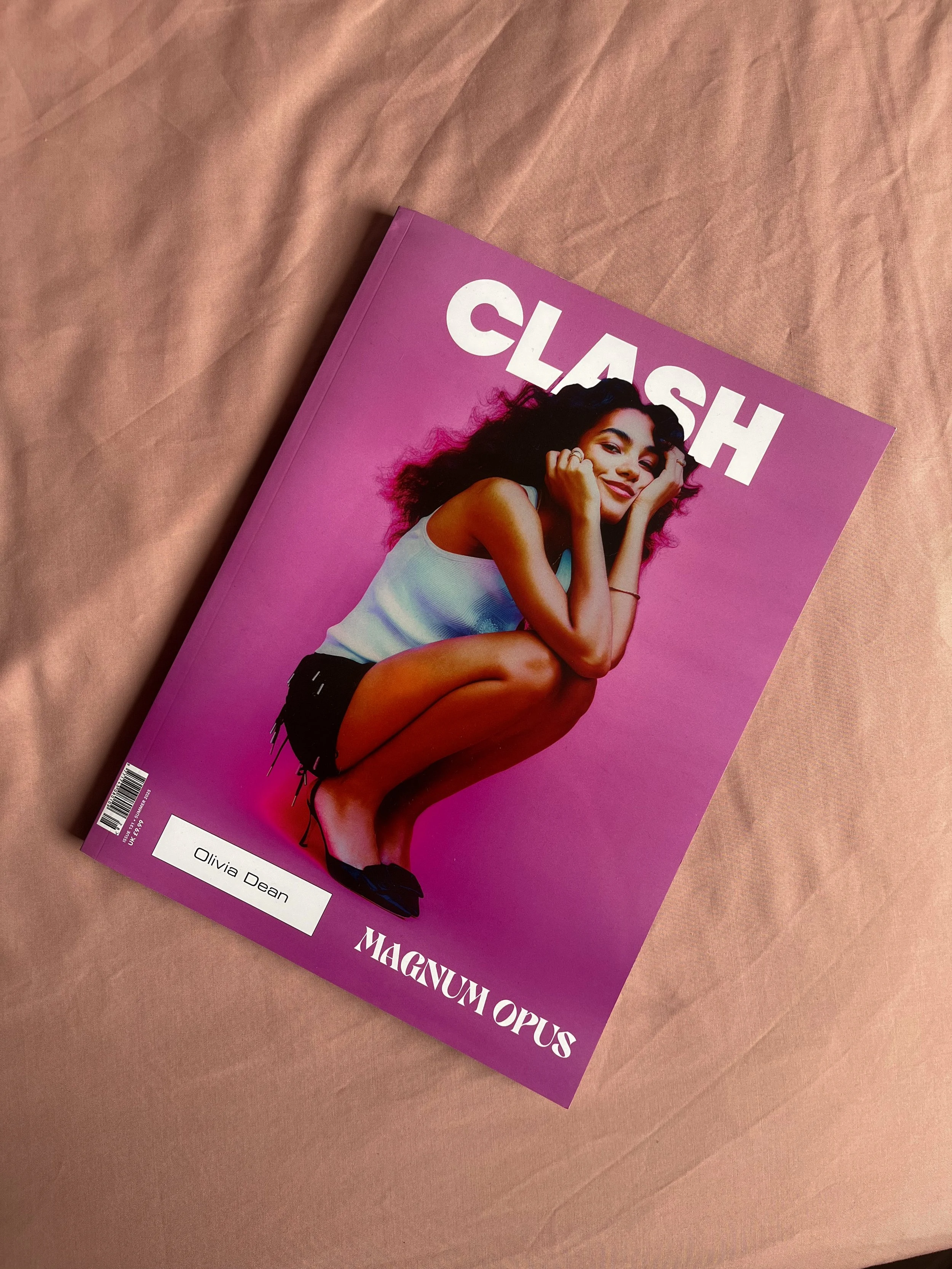 A magazine titled 'Clash' with a cover photo of a young woman squatting, smiling, with curly hair, wearing a white tank top, black shorts, and black shoes, set against a pink background.