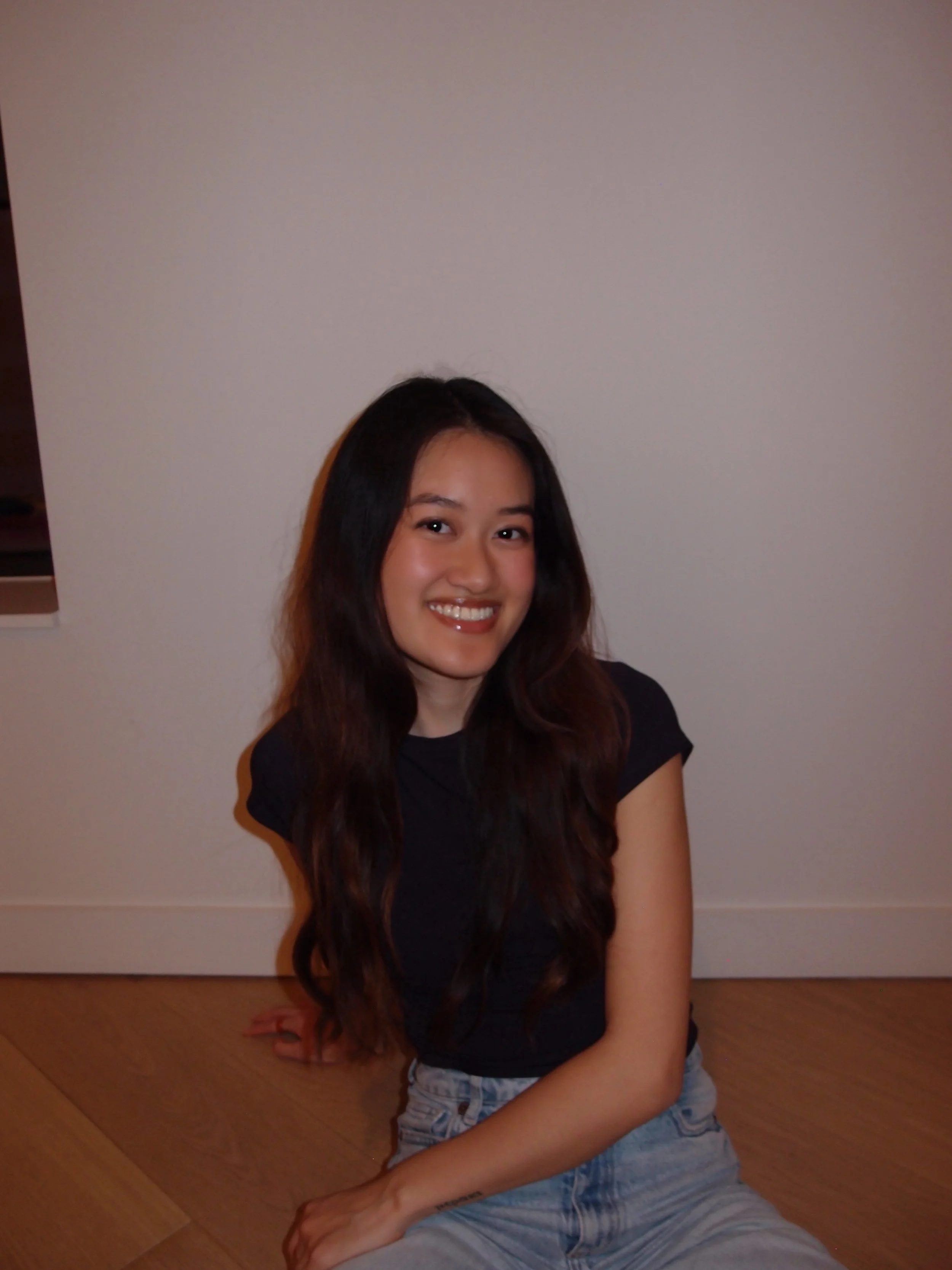 A young woman with long dark hair, smiling, sitting on a wooden floor against a plain white wall, wearing a black t-shirt and light blue jeans.