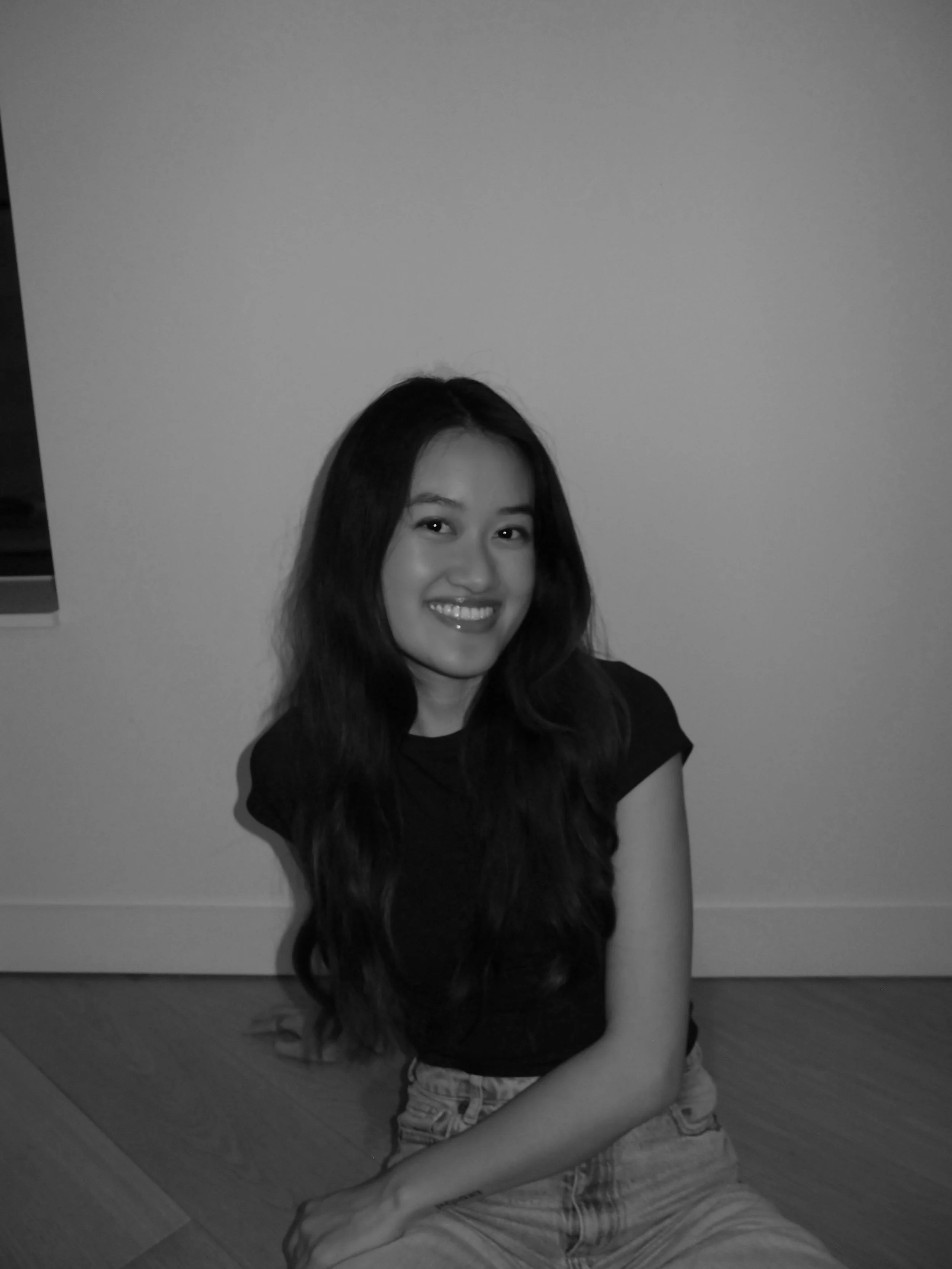 A woman smiling, sitting on the floor with long wavy hair, wearing a black top and light-colored jeans, in front of a plain wall.