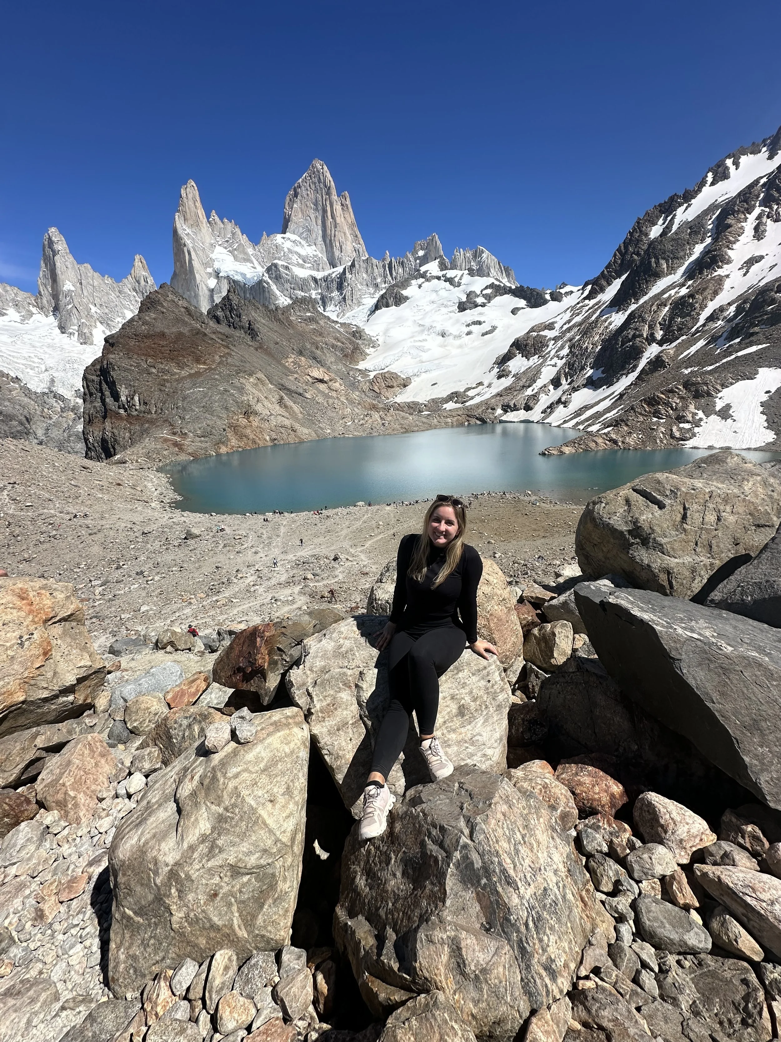 A woman sitting on rocks in a mountainous landscape with snowy peaks, a lake, and clear blue sky in El Chalten, Patagonia, Argentina.