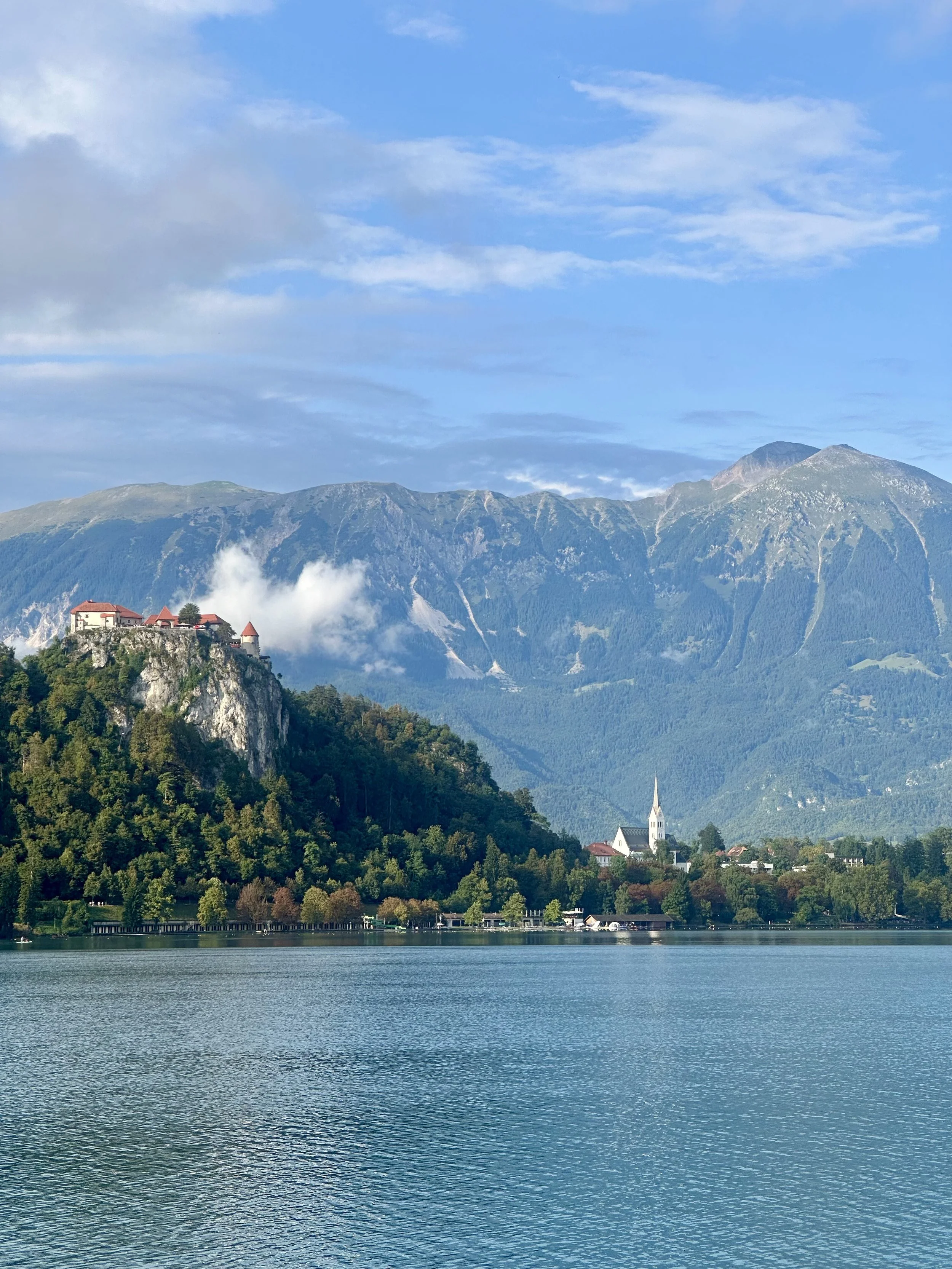 Scenic view of Bled Lake in Slovenia with a church and houses on the hillside, surrounded by green mountains and a partly cloudy sky.