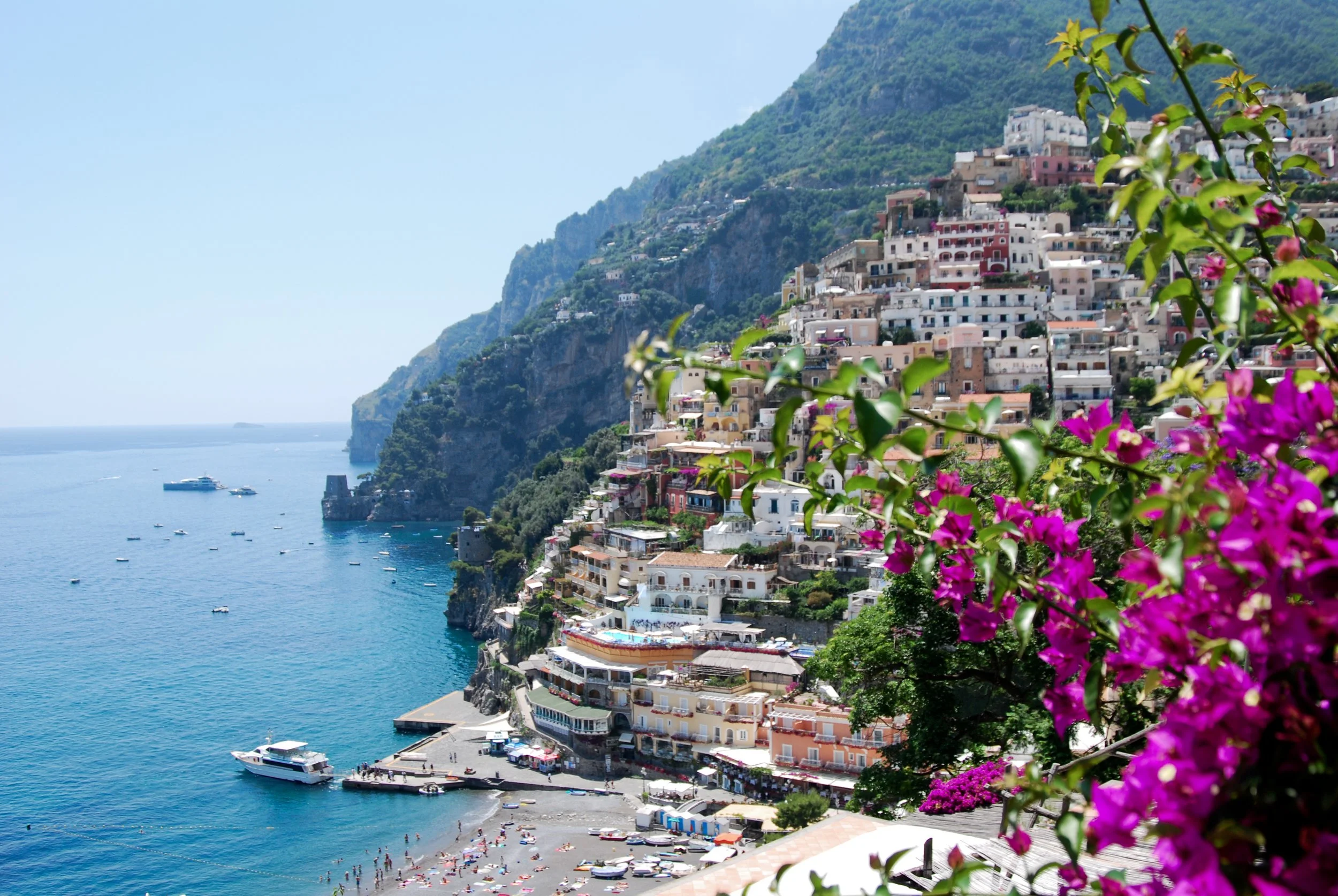 Positano Italy with colorful buildings on a hillside overlooking the sea, with boats docked and a beach with people swimming and sunbathing, framed by pink flowers in the foreground.