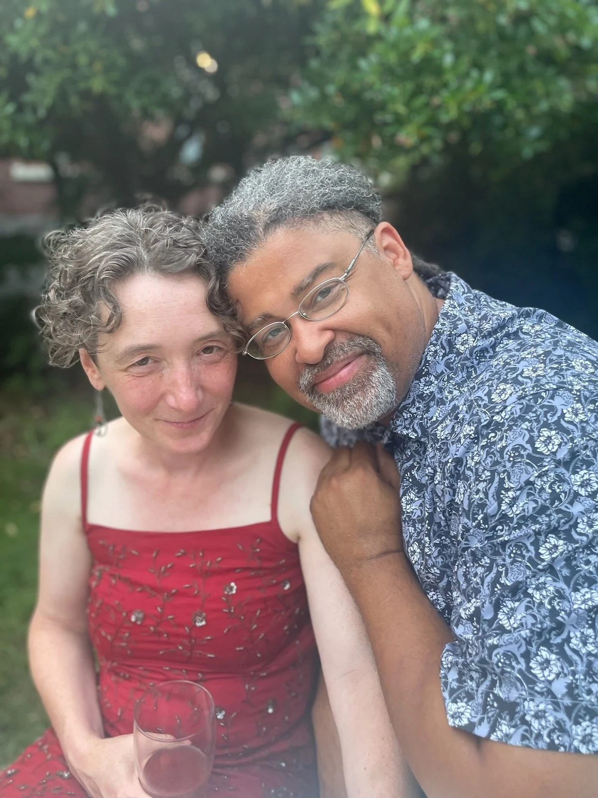 A woman with short curly gray hair wearing a red dress, and a man with glasses, gray hair, and a beard wearing a patterned shirt, smiling together outdoors.