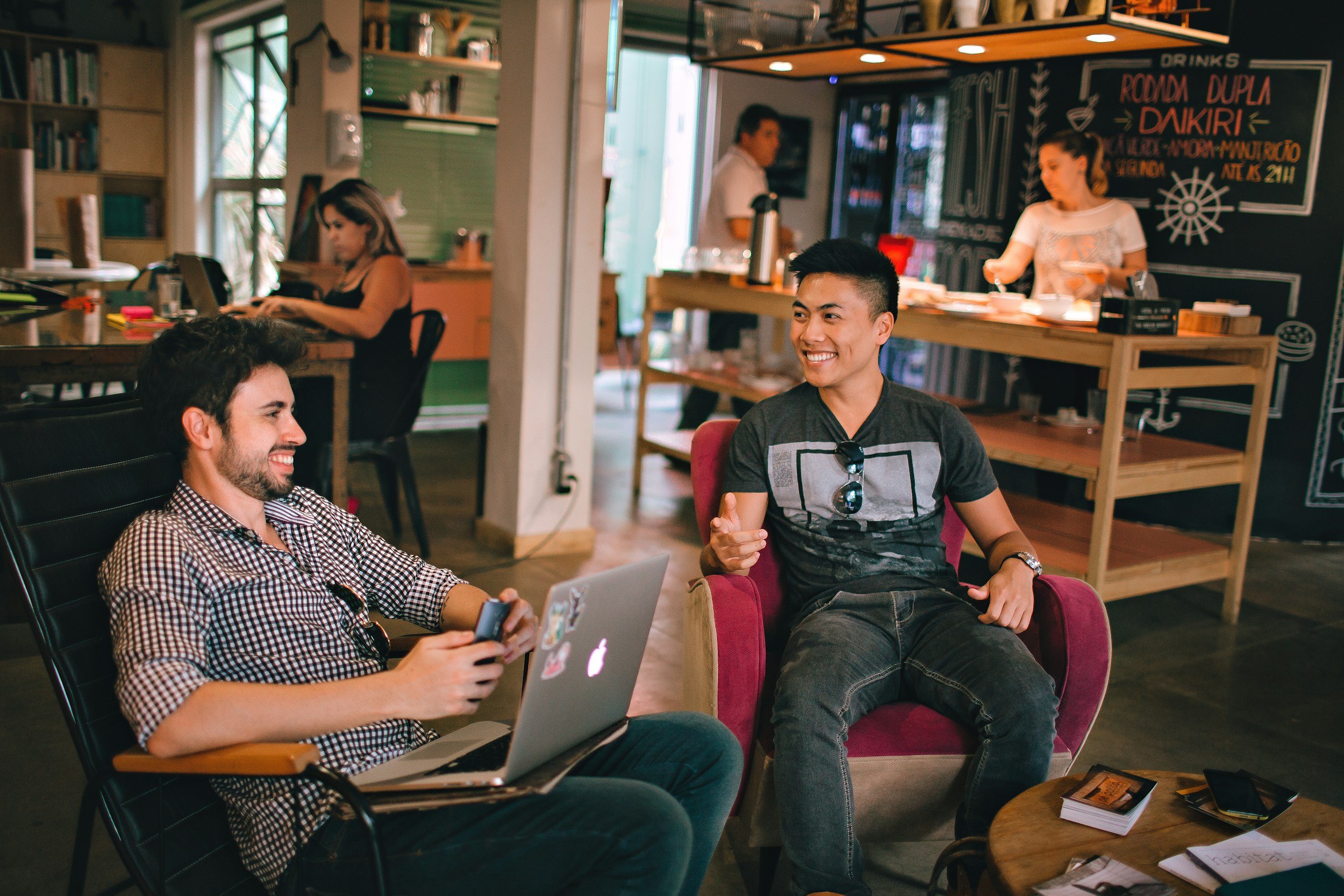 Zwei junge Männer lachen und unterhalten sich in einem gemütlichen Café. Einer sitzt mit einem Laptop und Smartphone, der andere sitzt in einem bequemen Sessel. Im Hintergrund sind weitere Café-Gäste und eine Bar mit Snacks und Getränken sichtbar.
