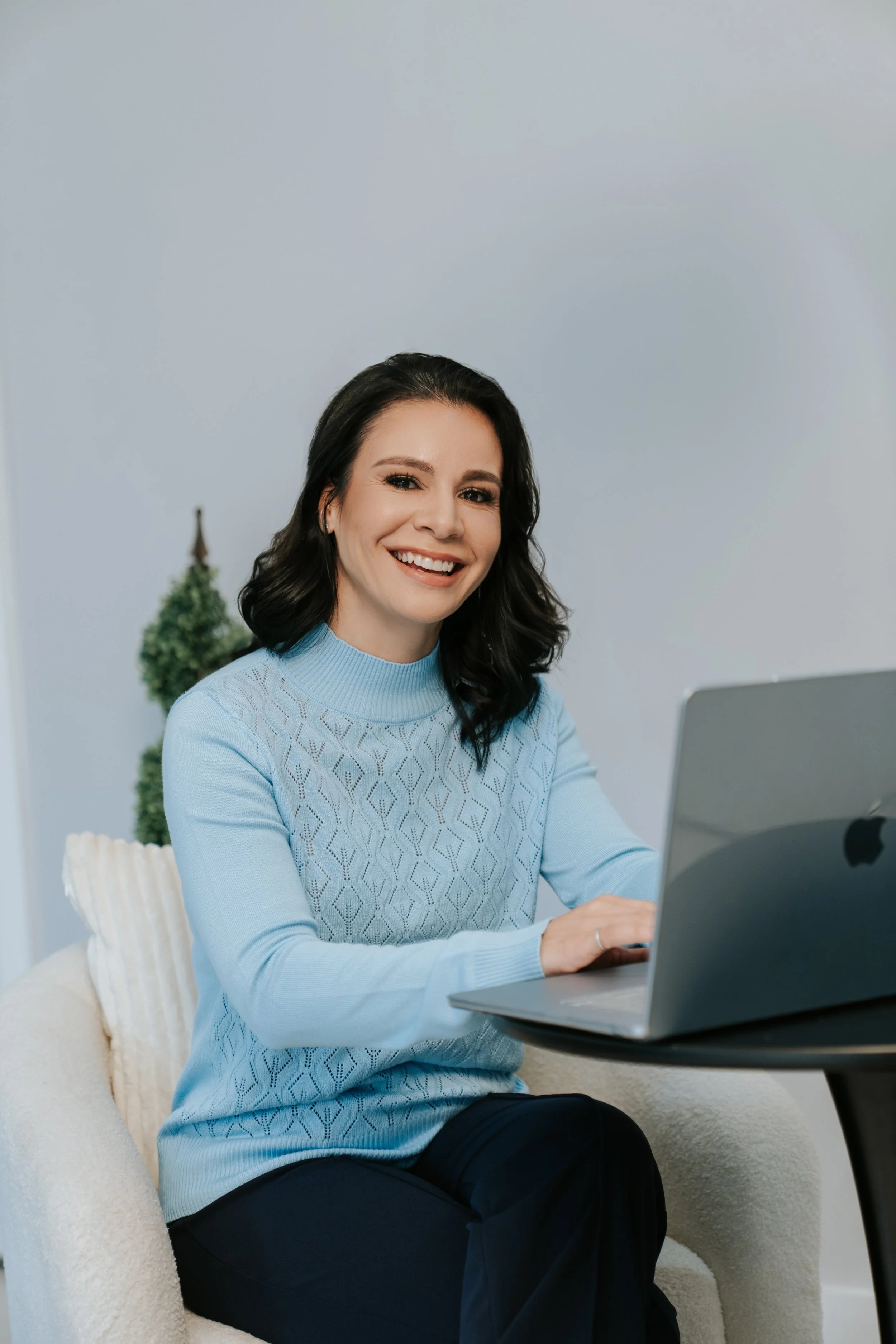 A smiling woman with dark hair wearing a light blue sweater sitting at a table using a laptop, with a decorated Christmas tree in the background.