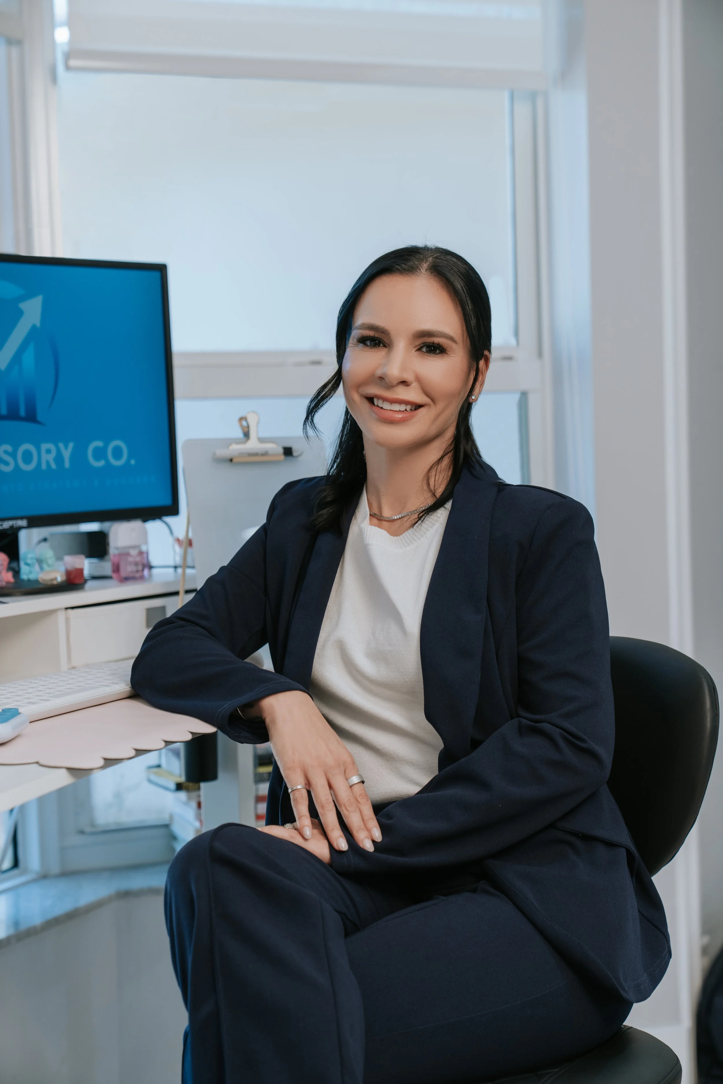 A woman in a professional setting sitting at a desk, smiling, with a computer monitor displaying a rising arrow and the text 'SORY CO.' behind her.