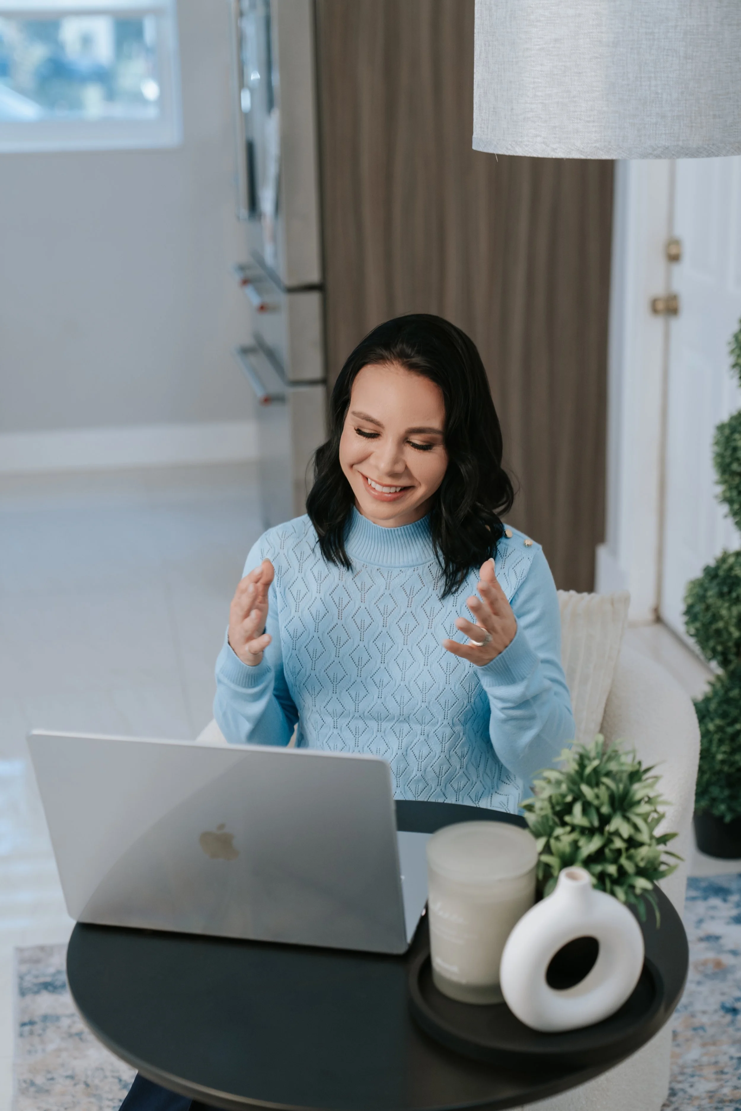 A woman with dark wavy hair, wearing a light blue sweater, sitting at a desk with a silver laptop, smiling with her hands near her face. In front of her on the desk are decorative white vases and a small green plant. The background includes a gray wall, a window, and a large lamp.