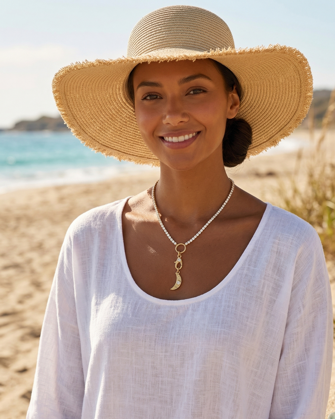 A woman smiling at the beach, wearing a large straw sun hat, white shirt, and a necklace with a crescent moon pendant.