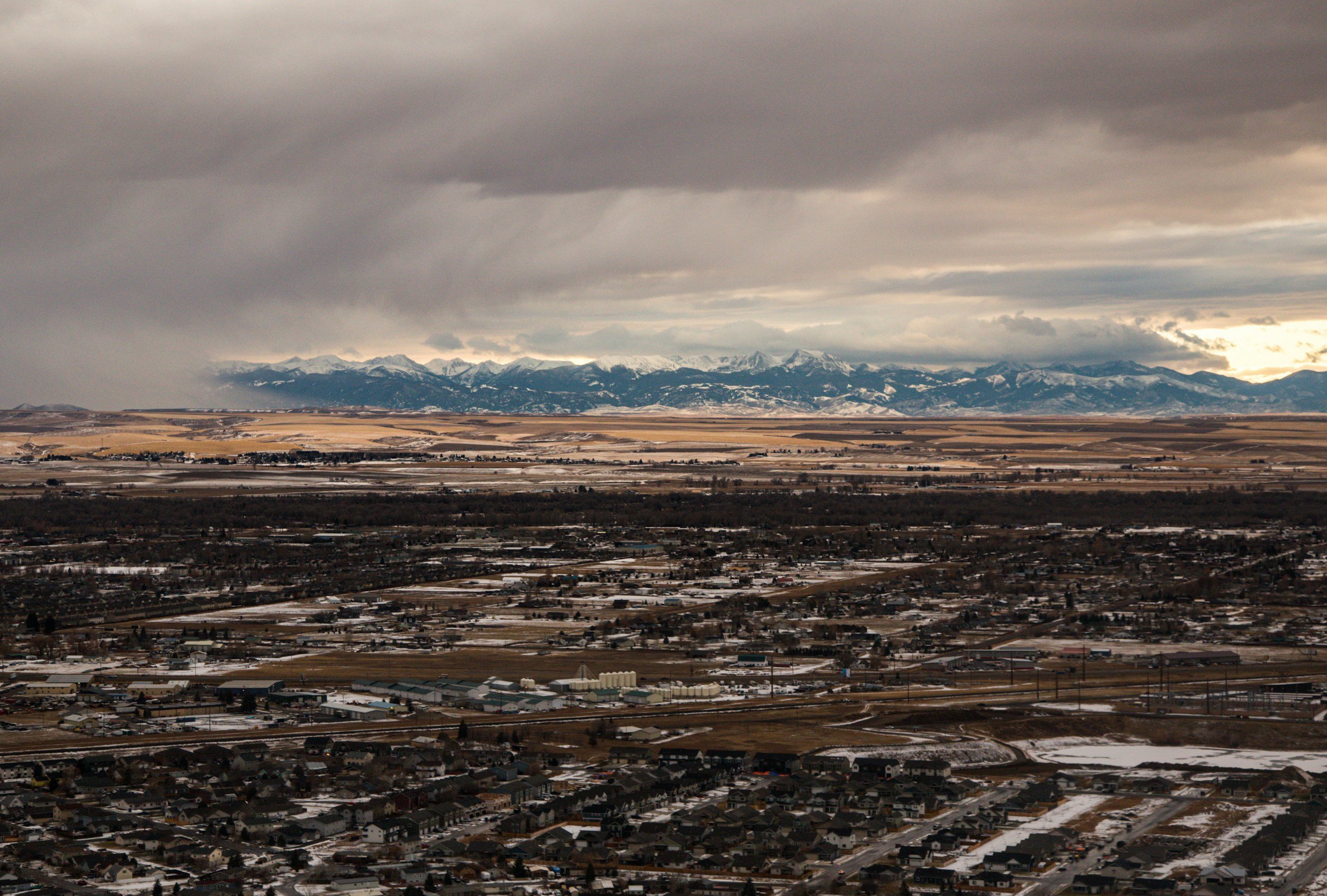 A landscape view of Bozeman Montana with houses and industrial buildings, snow patches on the ground, with mountains in the background under a cloudy sky.