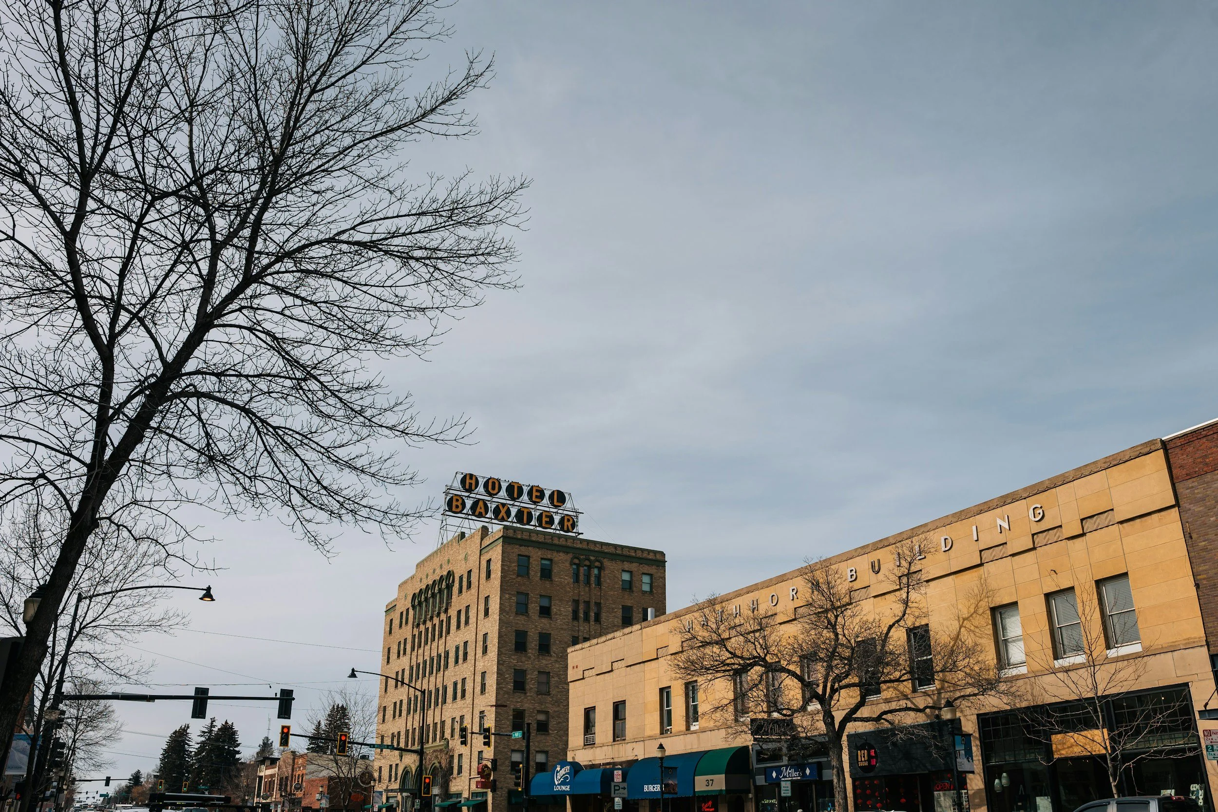 City street view with leafless trees, buildings, and a sign reading Hotel Baxter. Traffic lights and street lamps are visible.