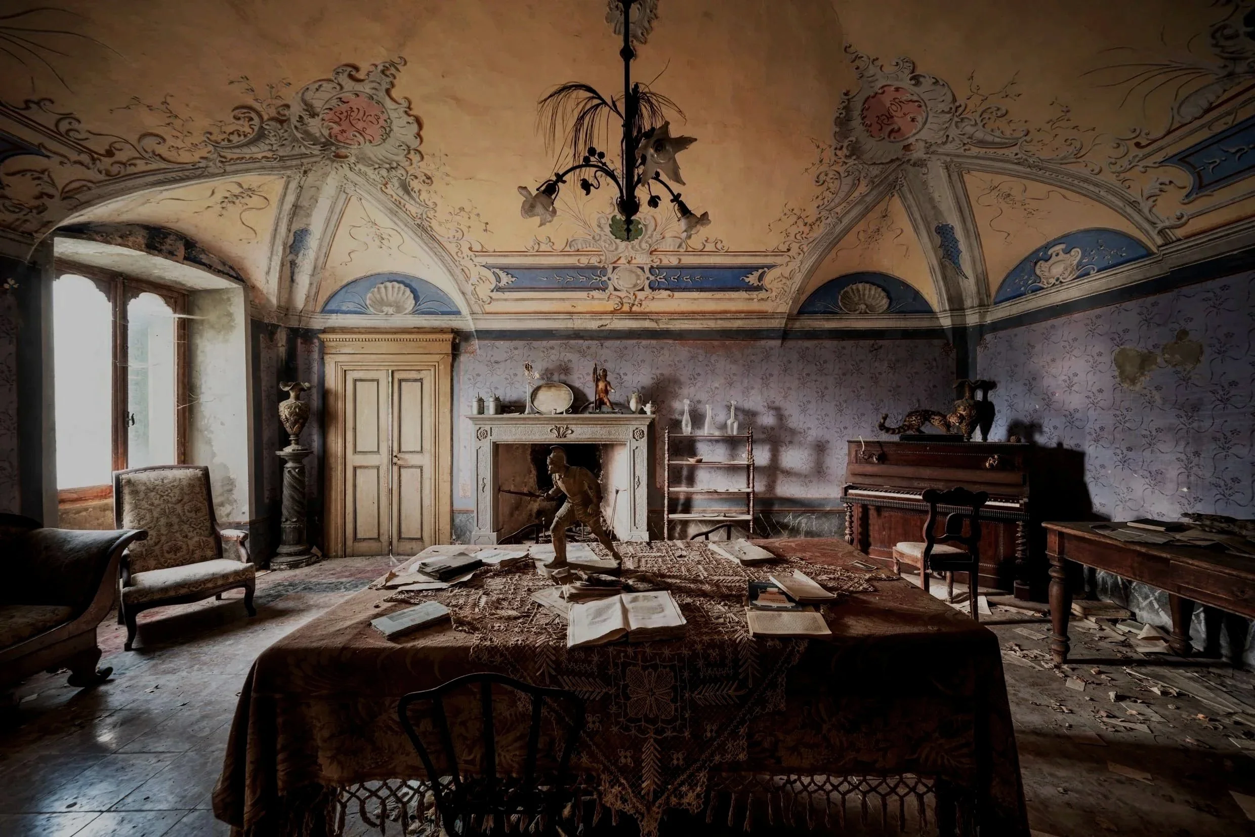 An old, abandoned room with ornate painted ceiling, fireplace, and vintage furniture. The room is messy with papers scattered on the table and dust on surfaces.