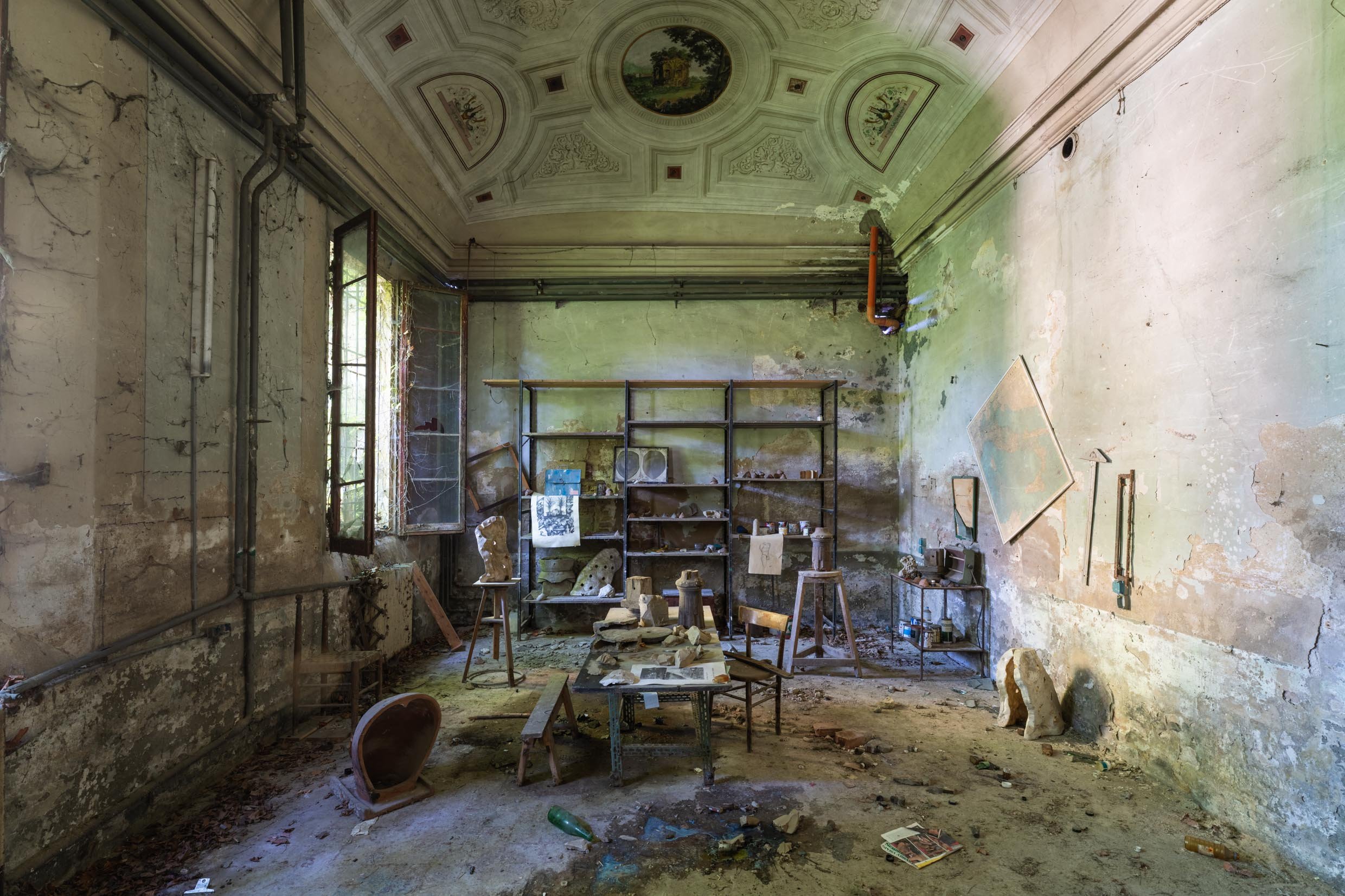 An abandoned room with peeling paint, broken furniture, and debris on the floor. The ceiling has ornate designs and paintings. There are open windows with overgrown plants outside, and sunlight is streaming in.