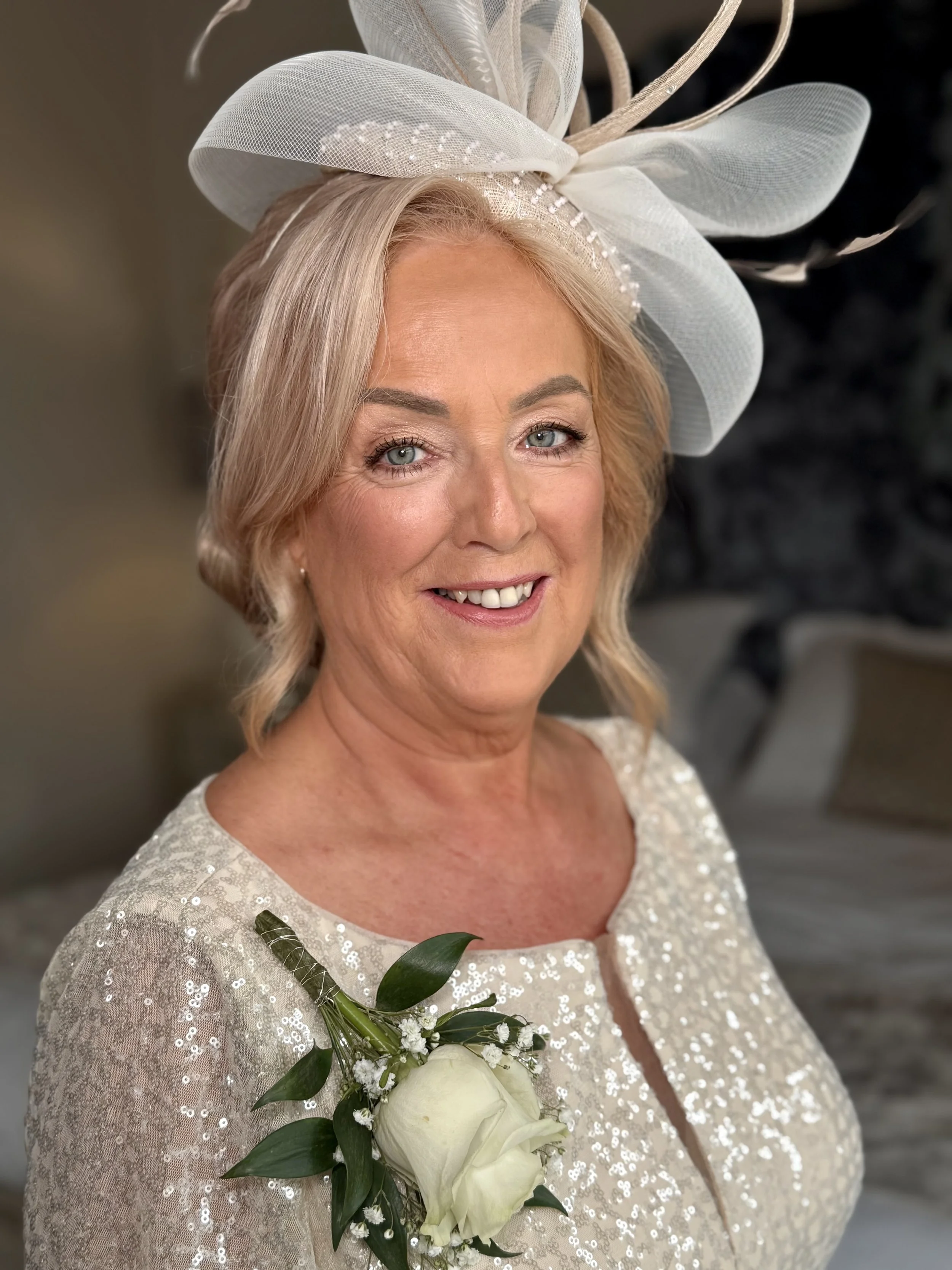 A woman wearing a large, white, fancy headpiece, smiling, holding a bouquet of white roses and greenery, dressed in a shimmery, light-colored dress with sequins, standing indoors.