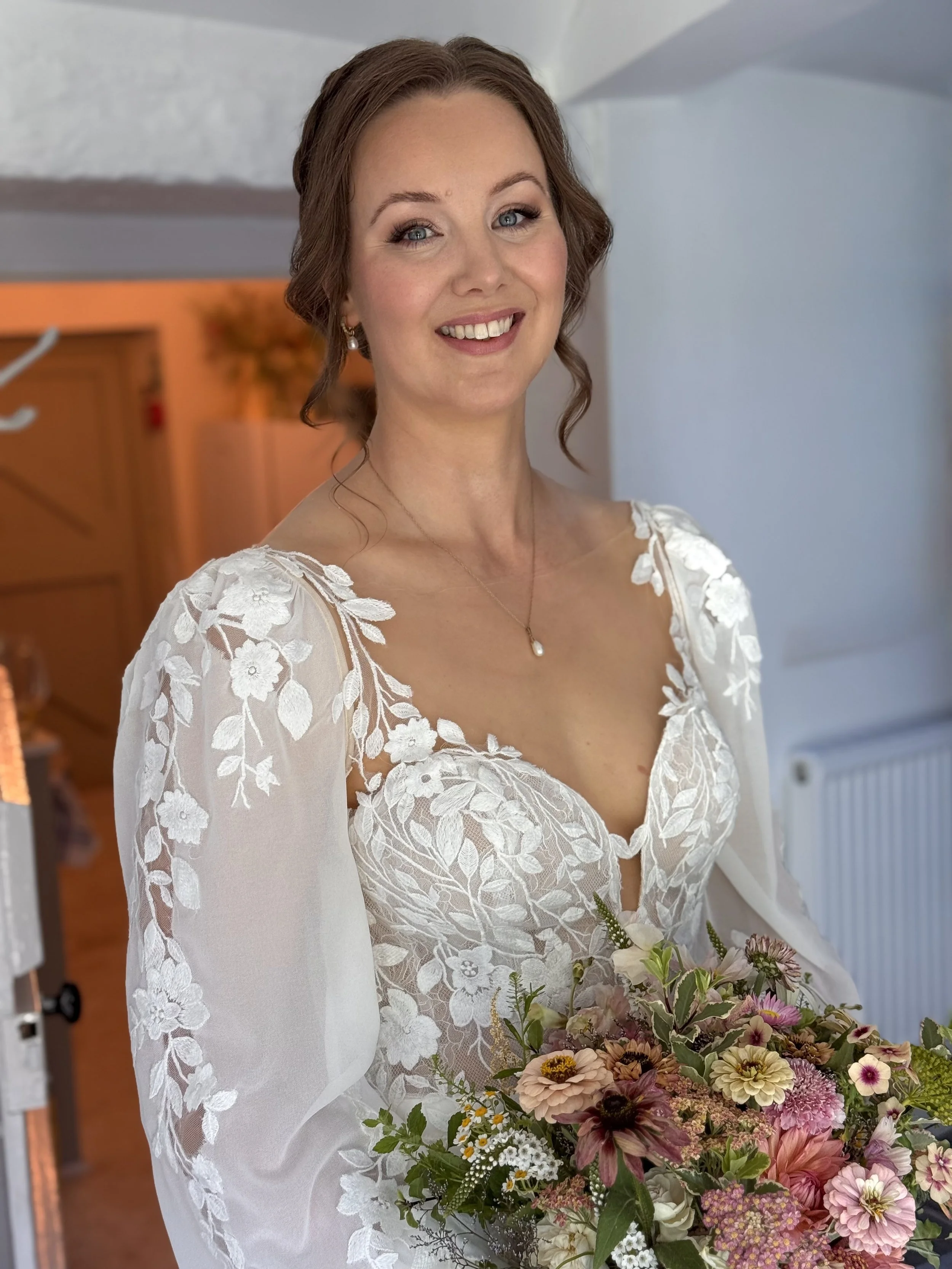 A woman in a white lace dress holding a colorful bouquet of flowers, smiling, with a blurred background.