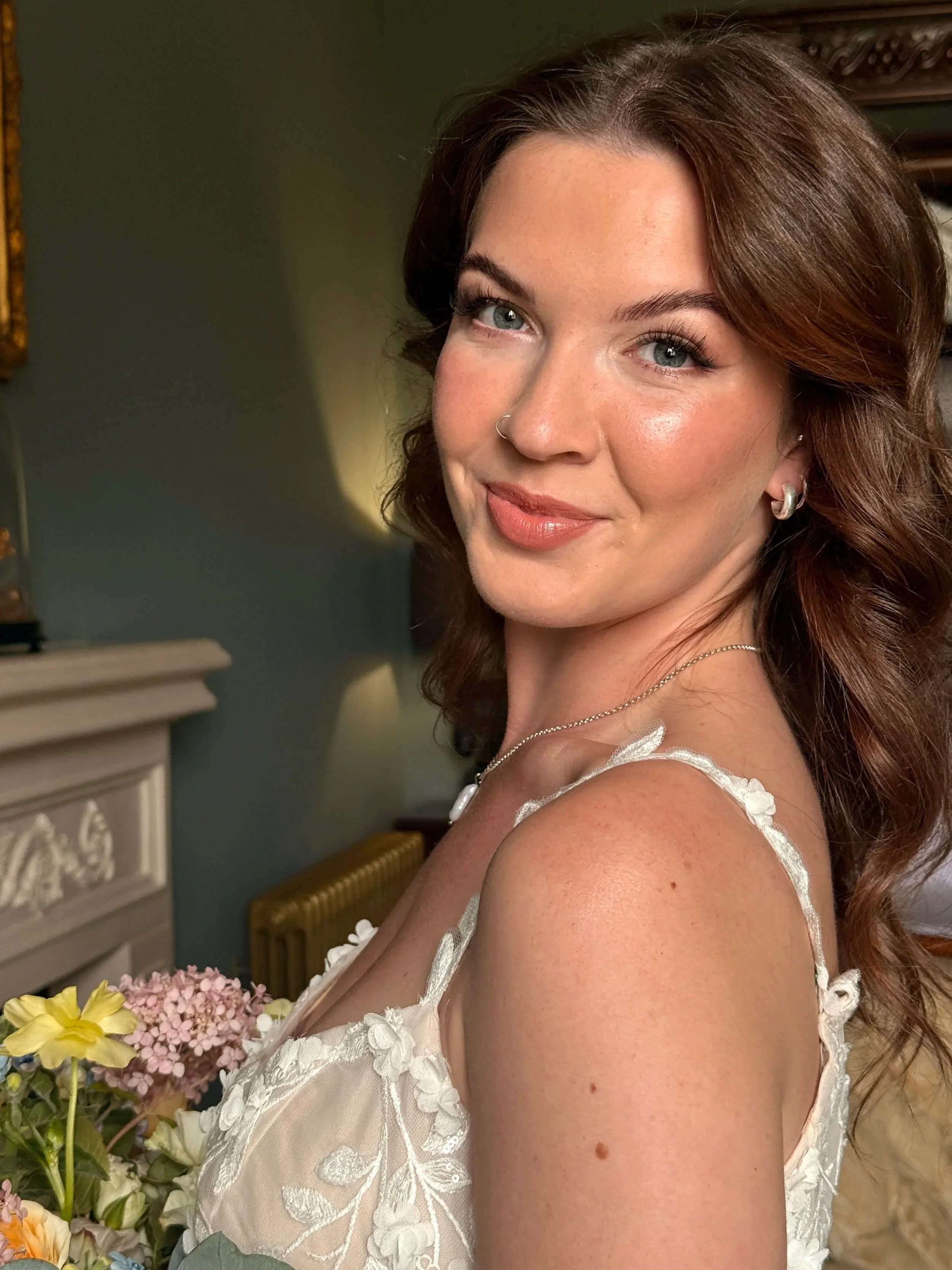 A woman with brown hair, blue eyes, and light skin wearing a white dress with floral embroidery, smiling, standing near a bouquet of pink and yellow flowers in a room with a fireplace and a mirror.