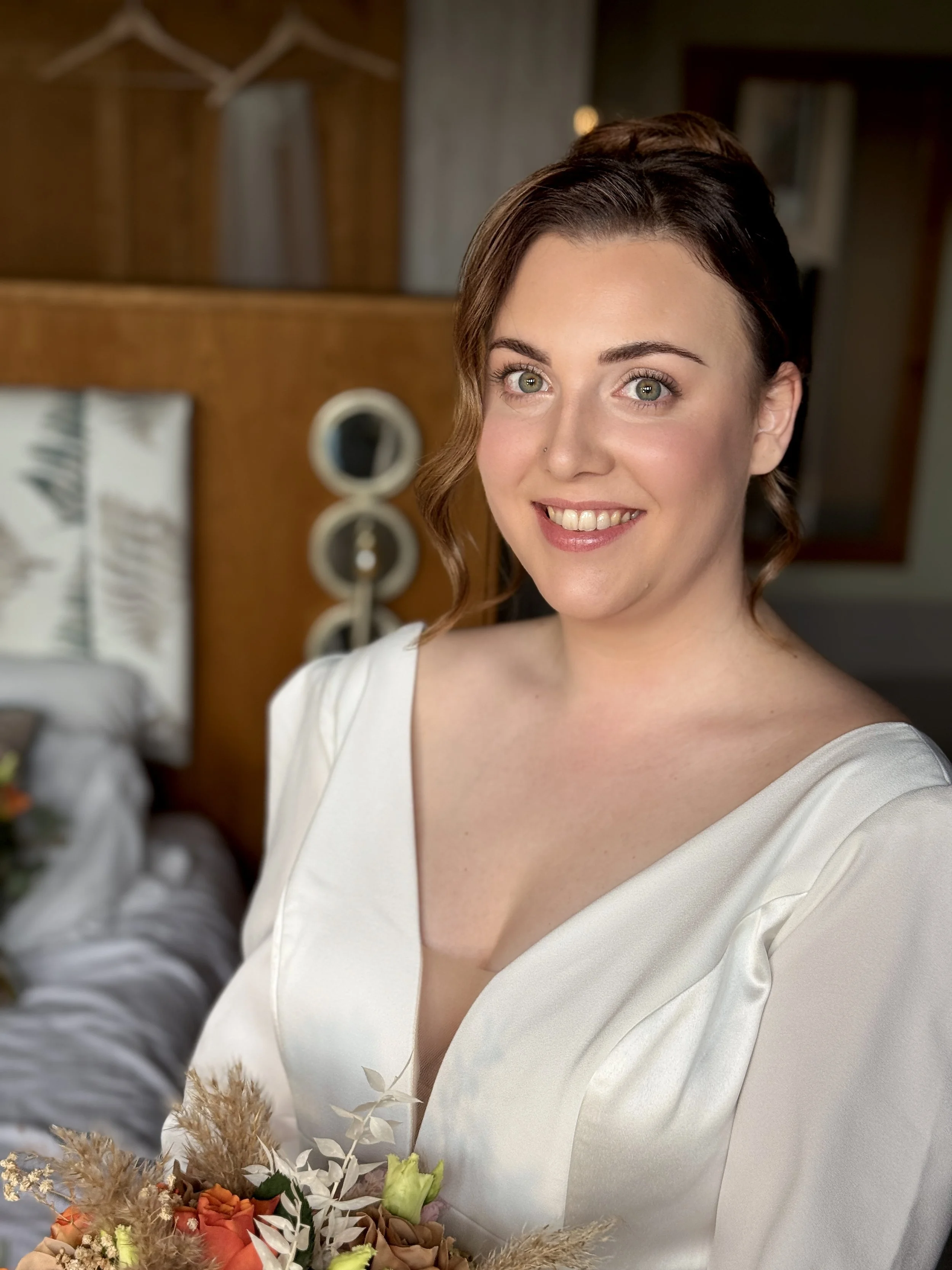 A smiling woman with short brown hair, wearing a white dress, holding a flower bouquet, standing indoors with a blurred background of a wooden shelf and decor.