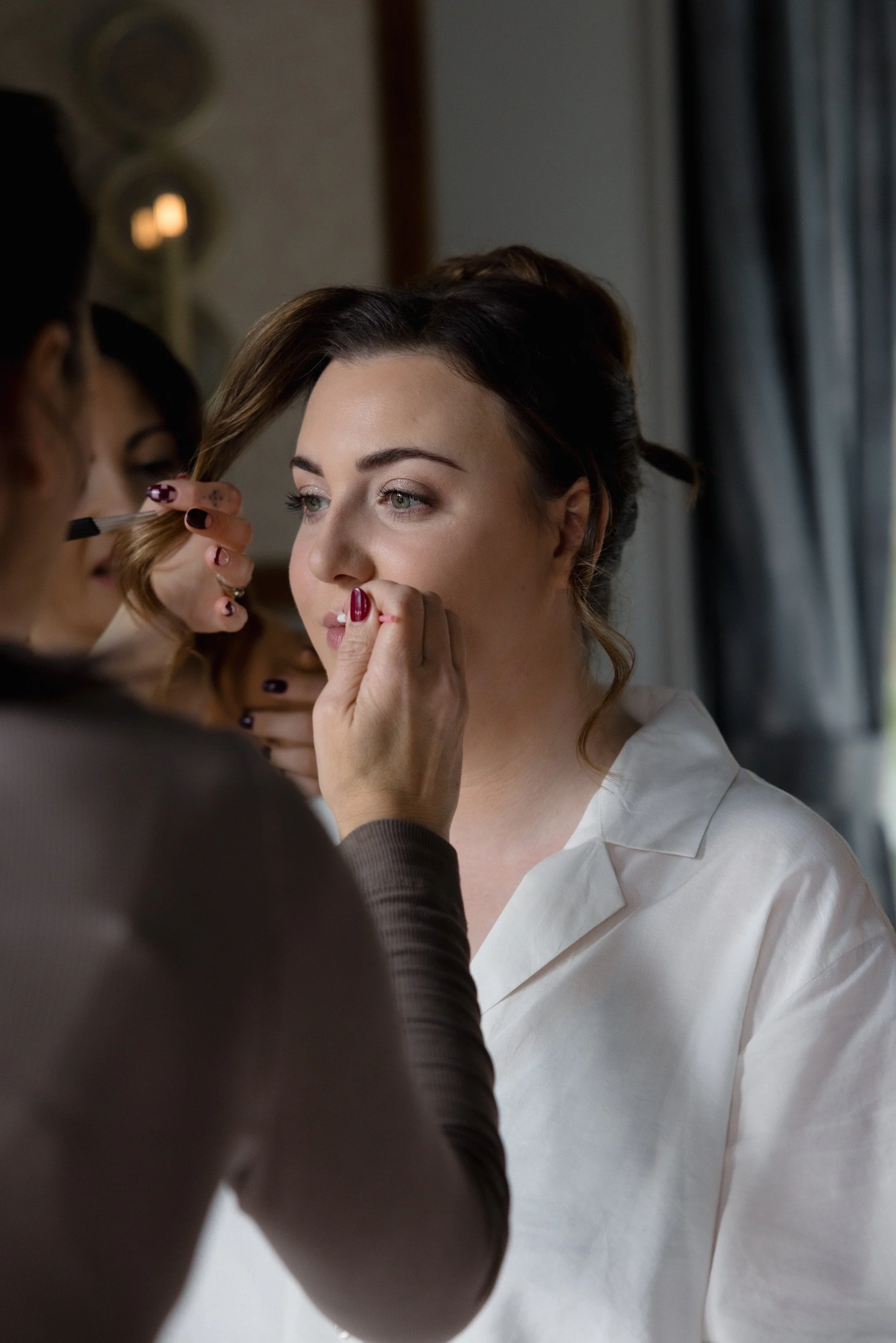 A woman with brown hair and light eyes receives makeup application from a makeup artist, with the artist applying makeup near her eye. The woman is wearing a white shirt, and the setting appears to be indoors with curtains and a mirror visible in the background.
