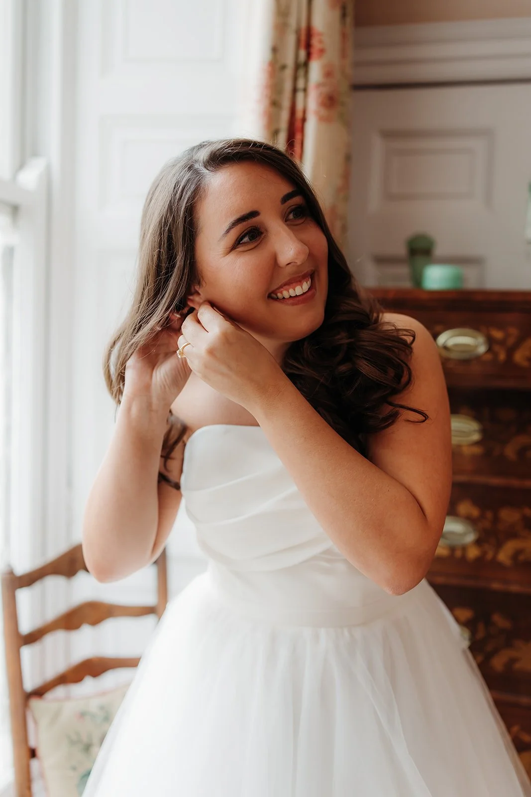 A smiling woman with long brown hair, wearing a white strapless dress, adjusting her earring near a window with floral curtains in a bright room.