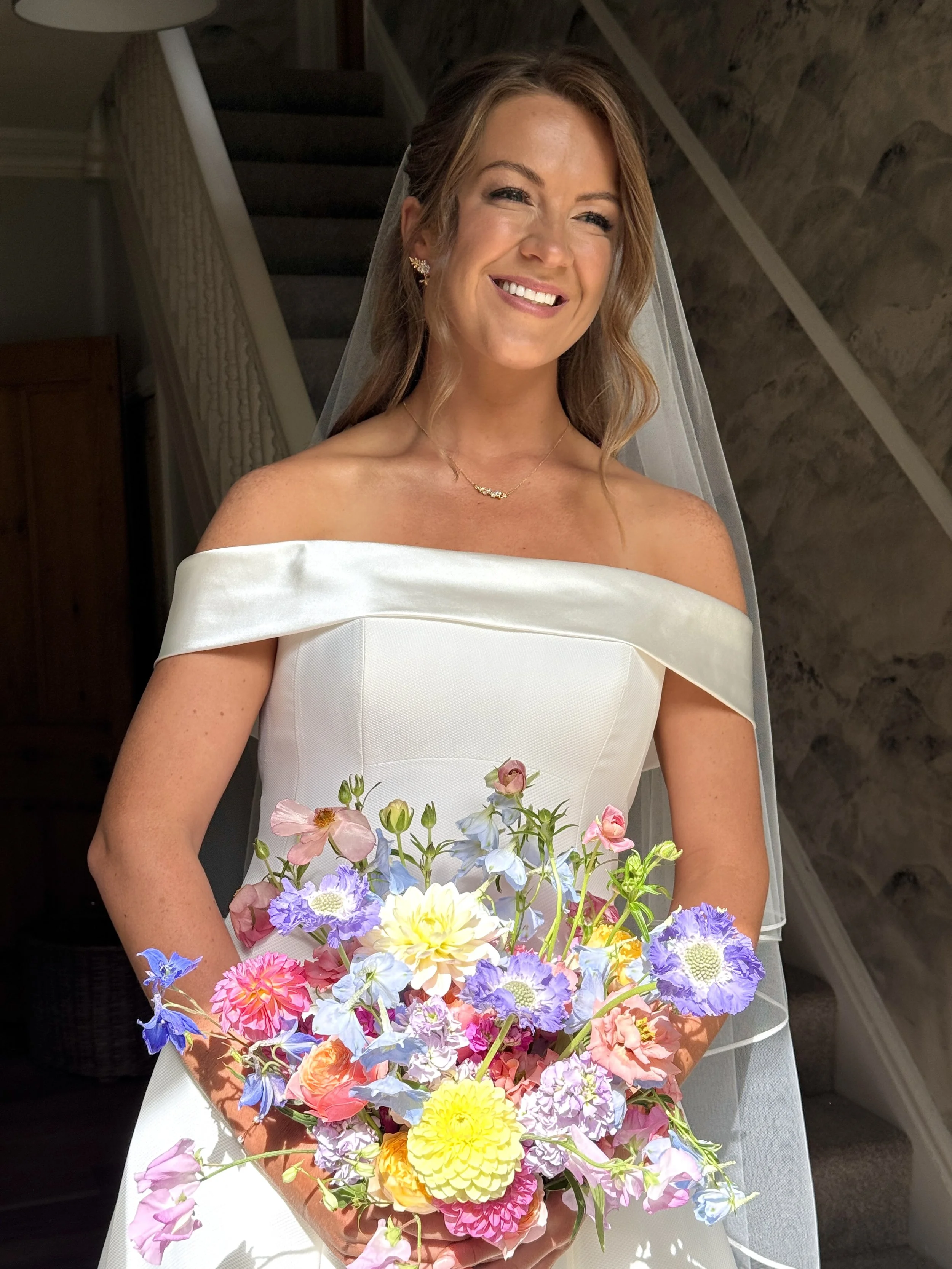 A smiling woman in a white off-the-shoulder dress holding a large colorful bouquet of flowers.