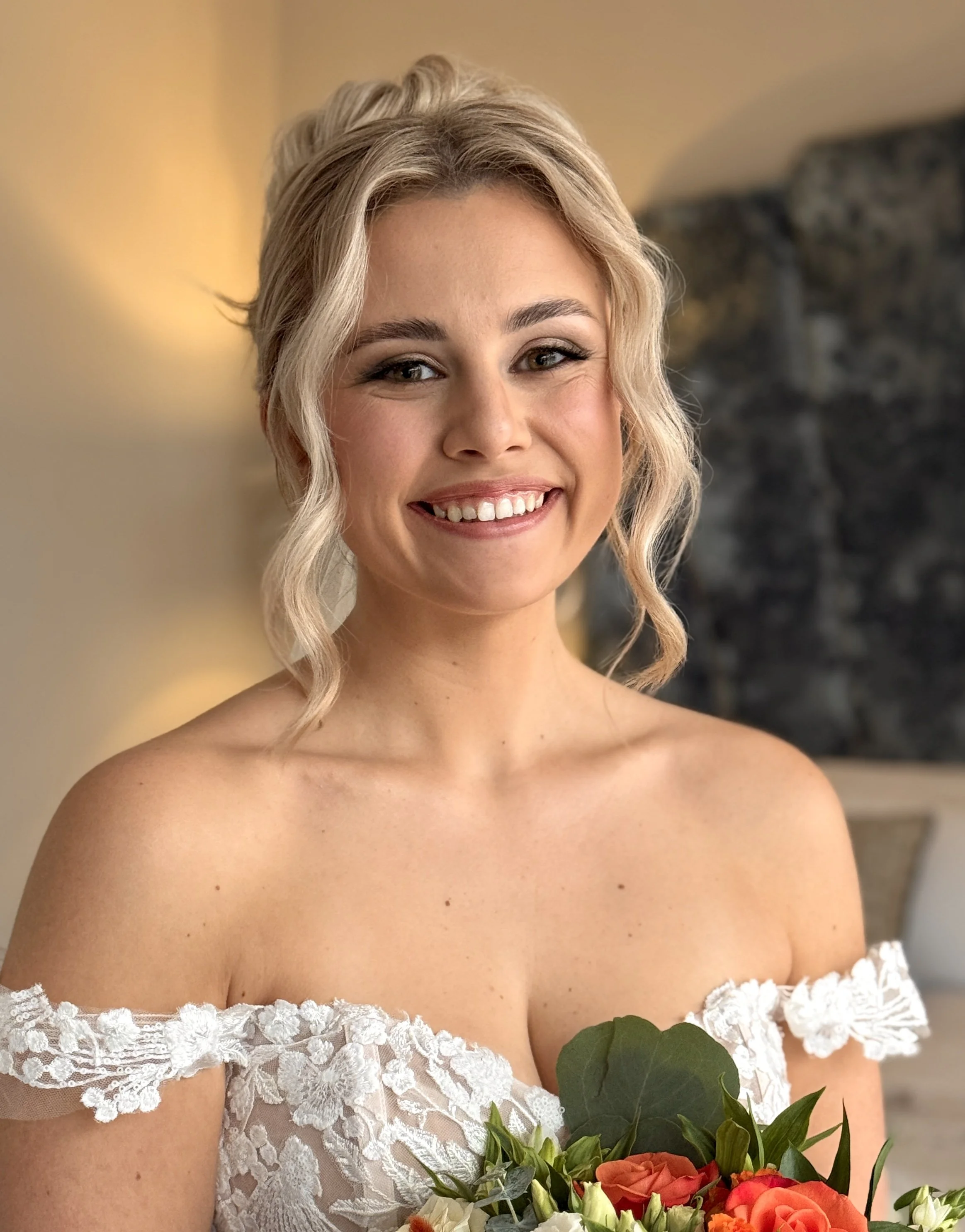 A smiling woman with blonde hair wearing a white lace off-the-shoulder dress, holding a bouquet of flowers with orange roses and green leaves, with a blurred background.