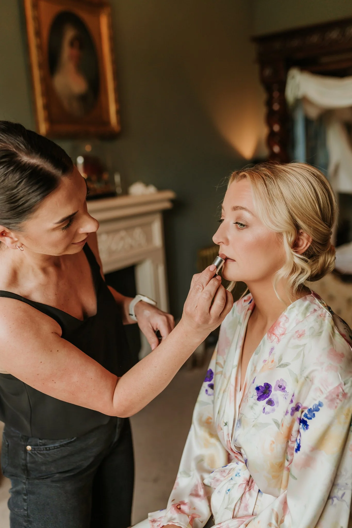 A woman in a floral robe is getting her makeup done by another woman in a black top in a cozy room with framed artwork and a fireplace in the background.