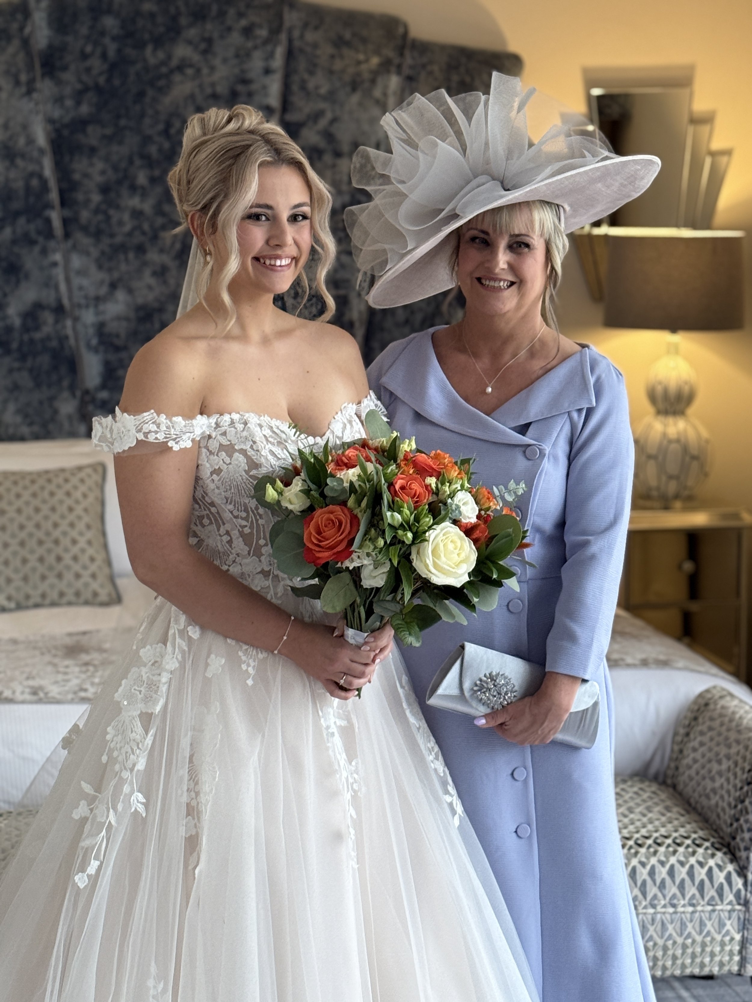 A young woman in a wedding dress holding a bouquet of flowers stands next to a woman in a light blue dress with a large, elaborate white hat. They are smiling indoors, with a bed, lampshade, and patterned pillows in the background.