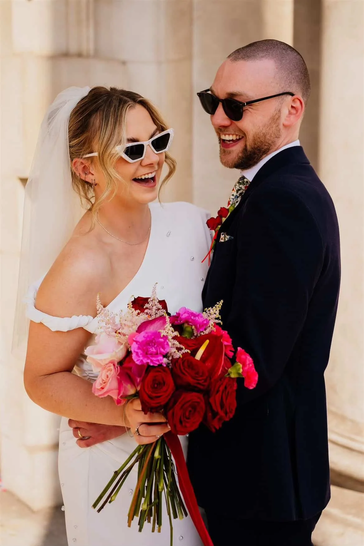 A bride and groom smiling and wearing sunglasses, holding a bouquet of pink and red flowers during a wedding ceremony.