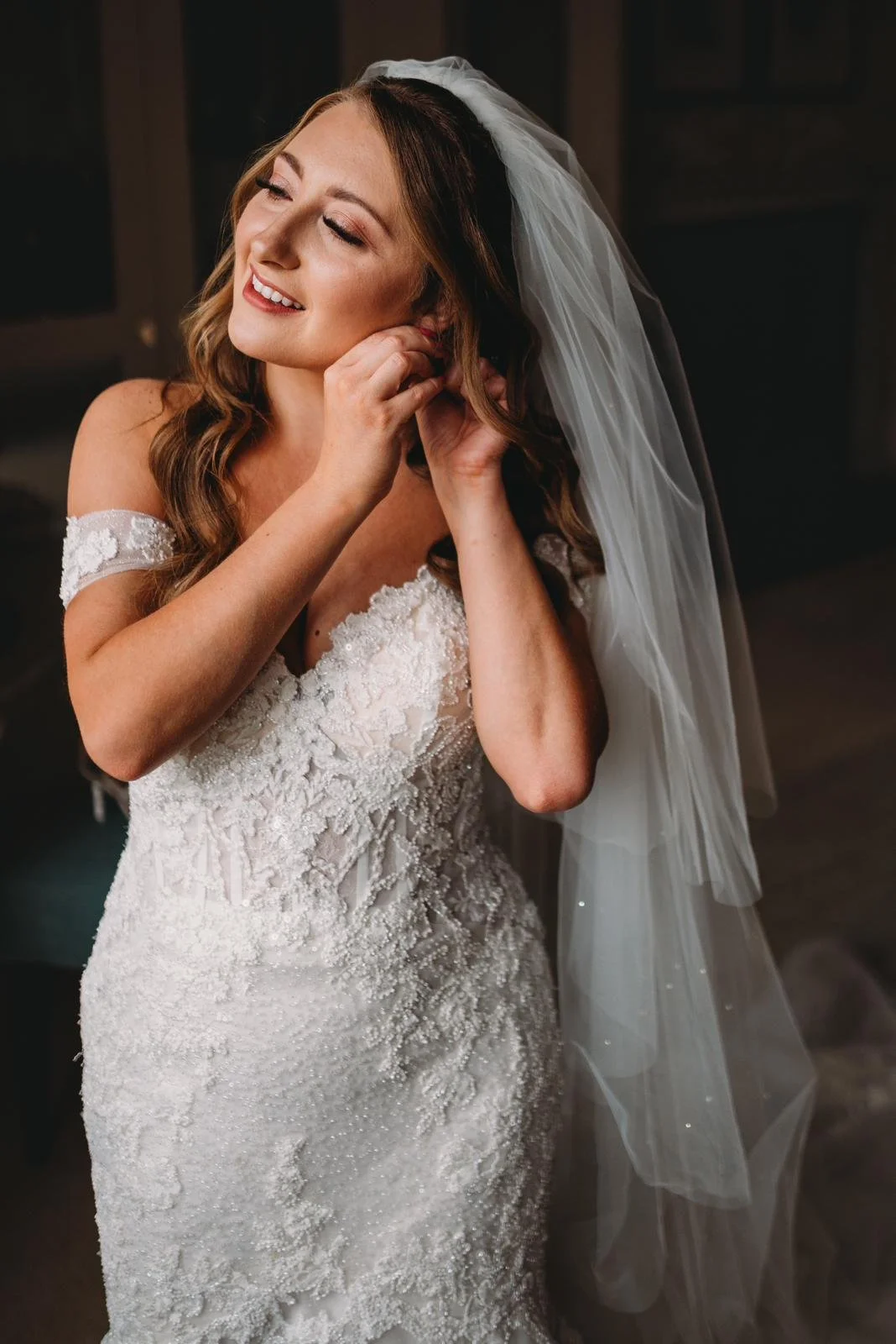 A woman in a wedding dress is putting on earrings with a smile.