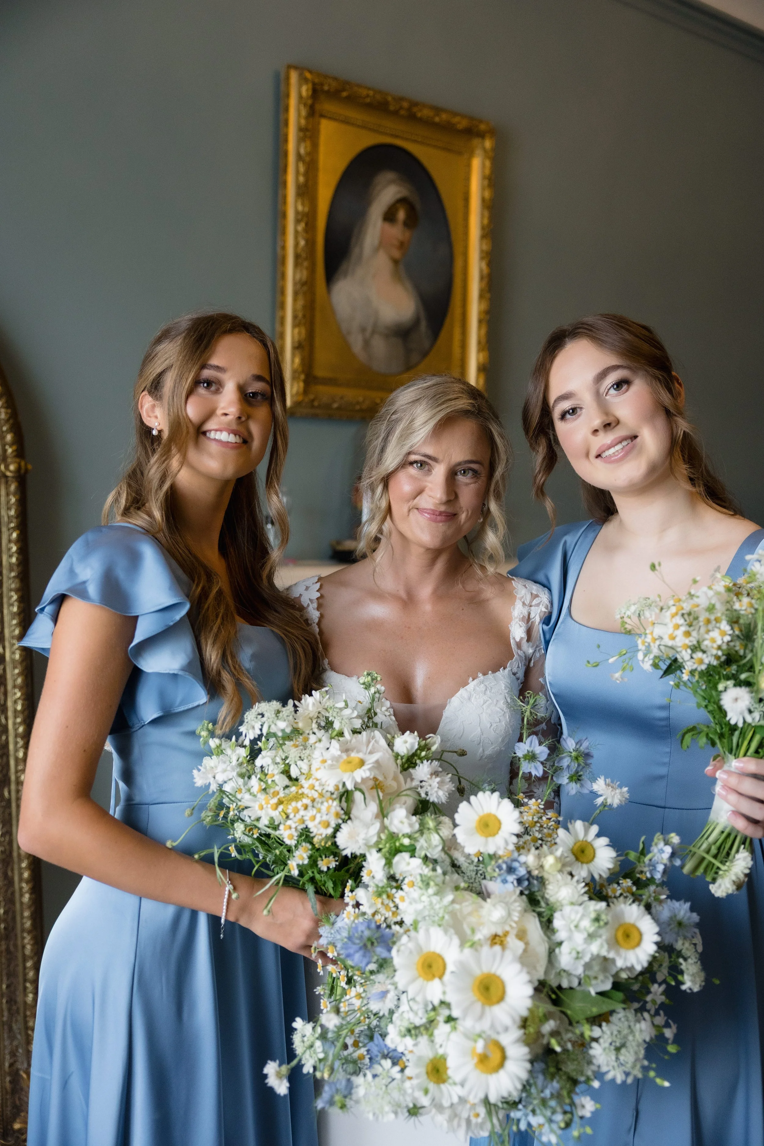 A bride and two bridesmaids at a wedding, holding bouquets of white and yellow flowers, standing in front of a portrait of a woman in a frame.