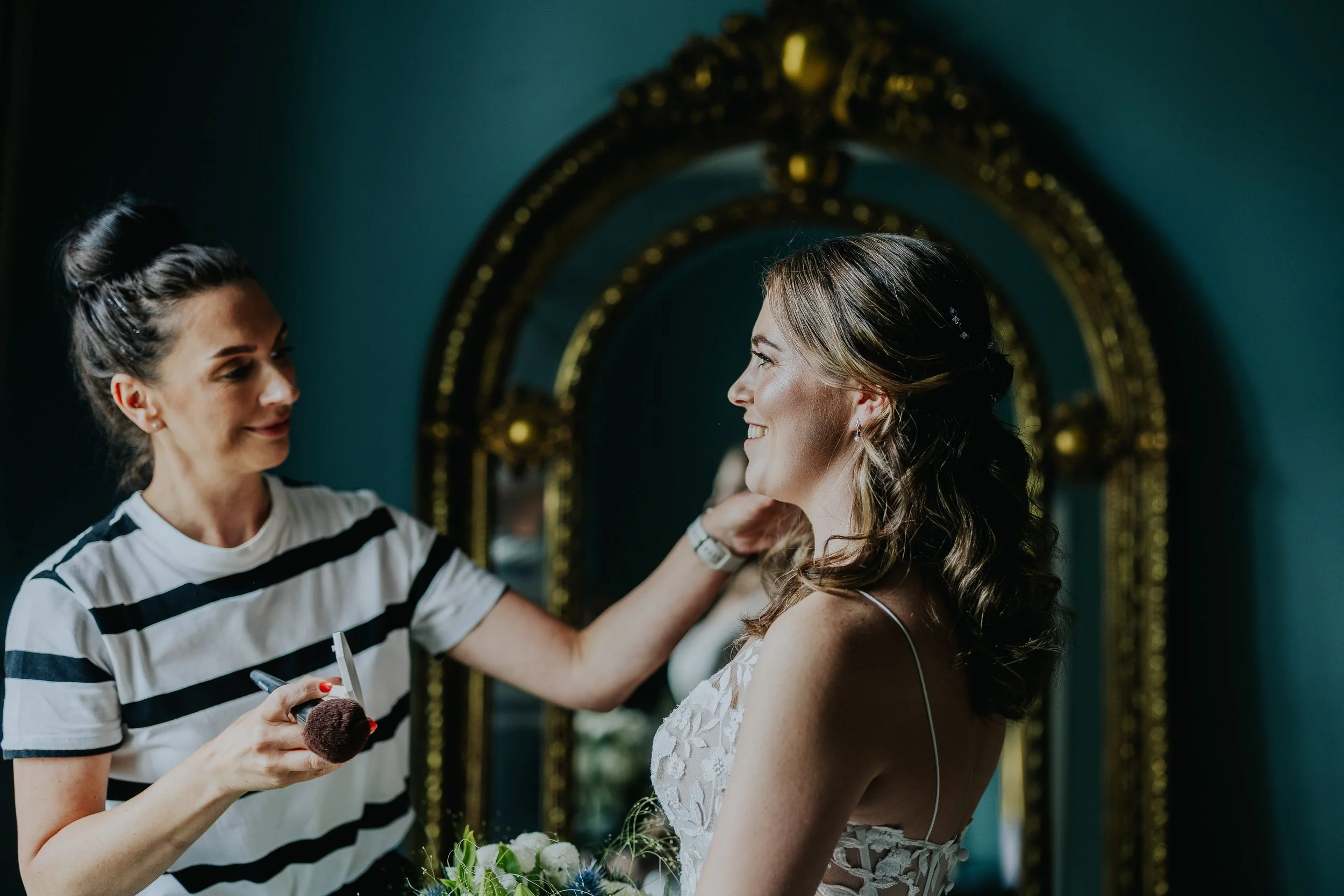 A woman in a white dress with lace details smiling as a makeup artist touches up her hair in front of a mirror in a fitting room.
