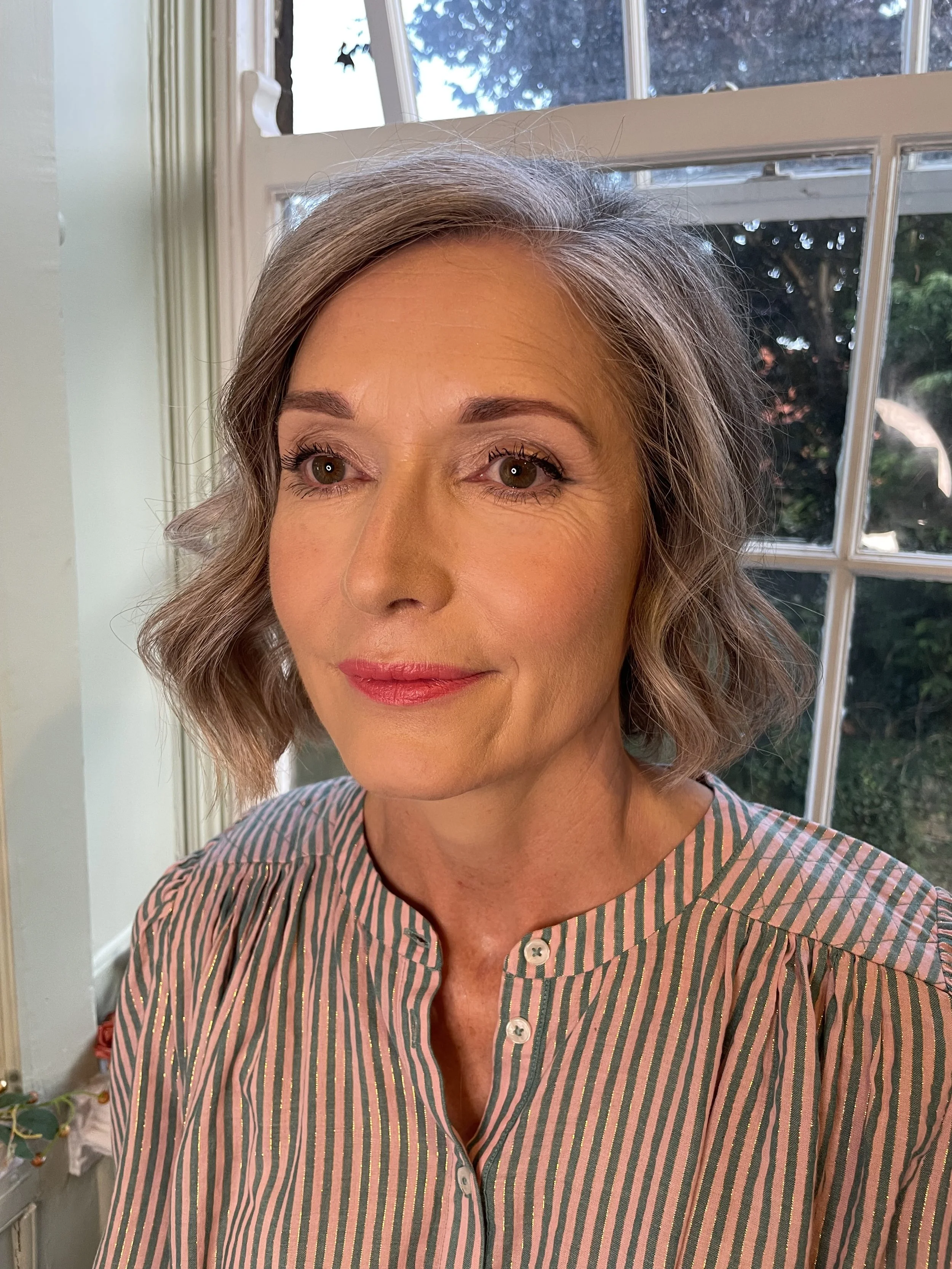 A middle-aged woman with shoulder-length gray hair, wearing a striped button-up shirt, standing near a window with green trees outside.