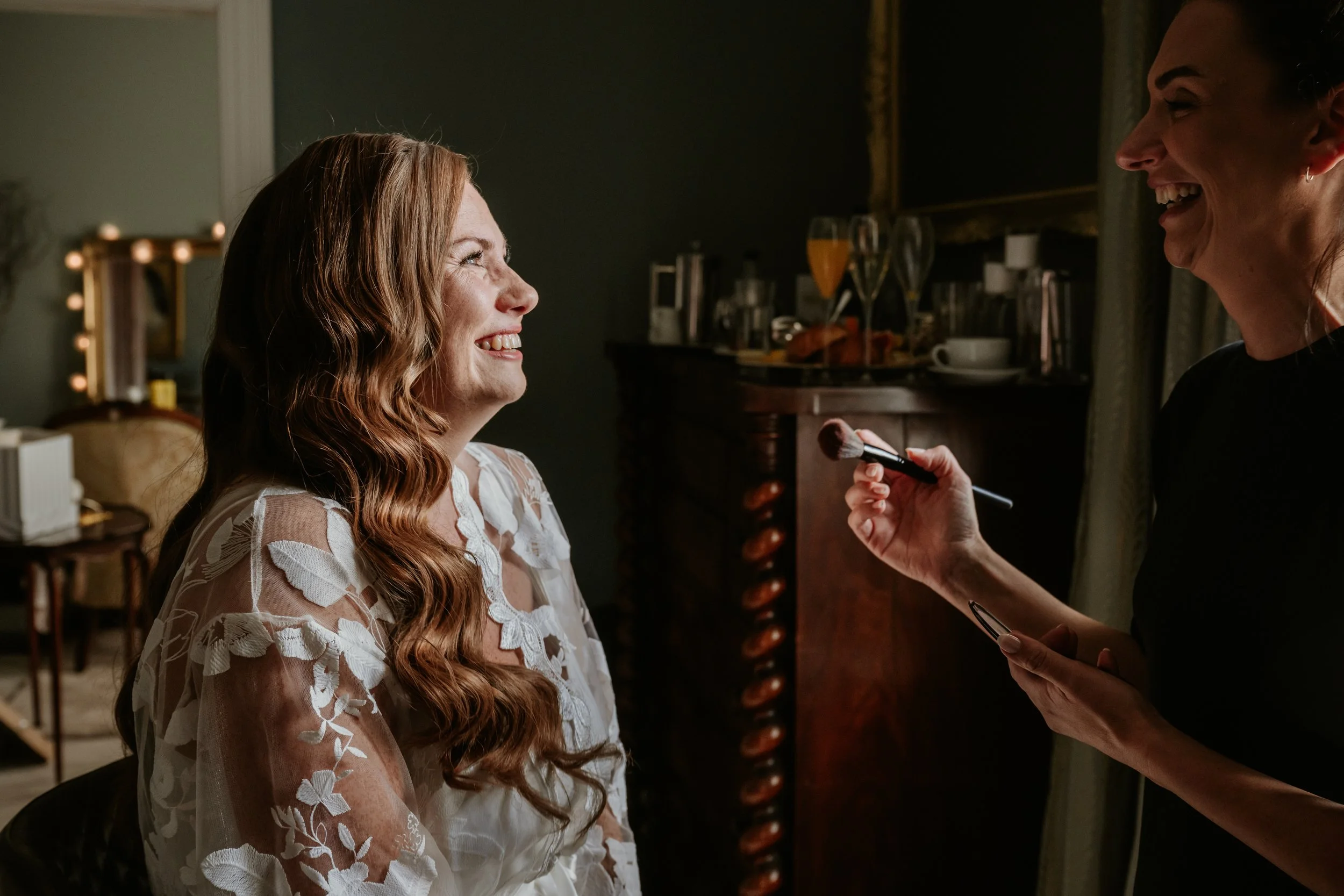 A woman with long red hair smiling as she gets her makeup done by another woman in a dimly lit room.