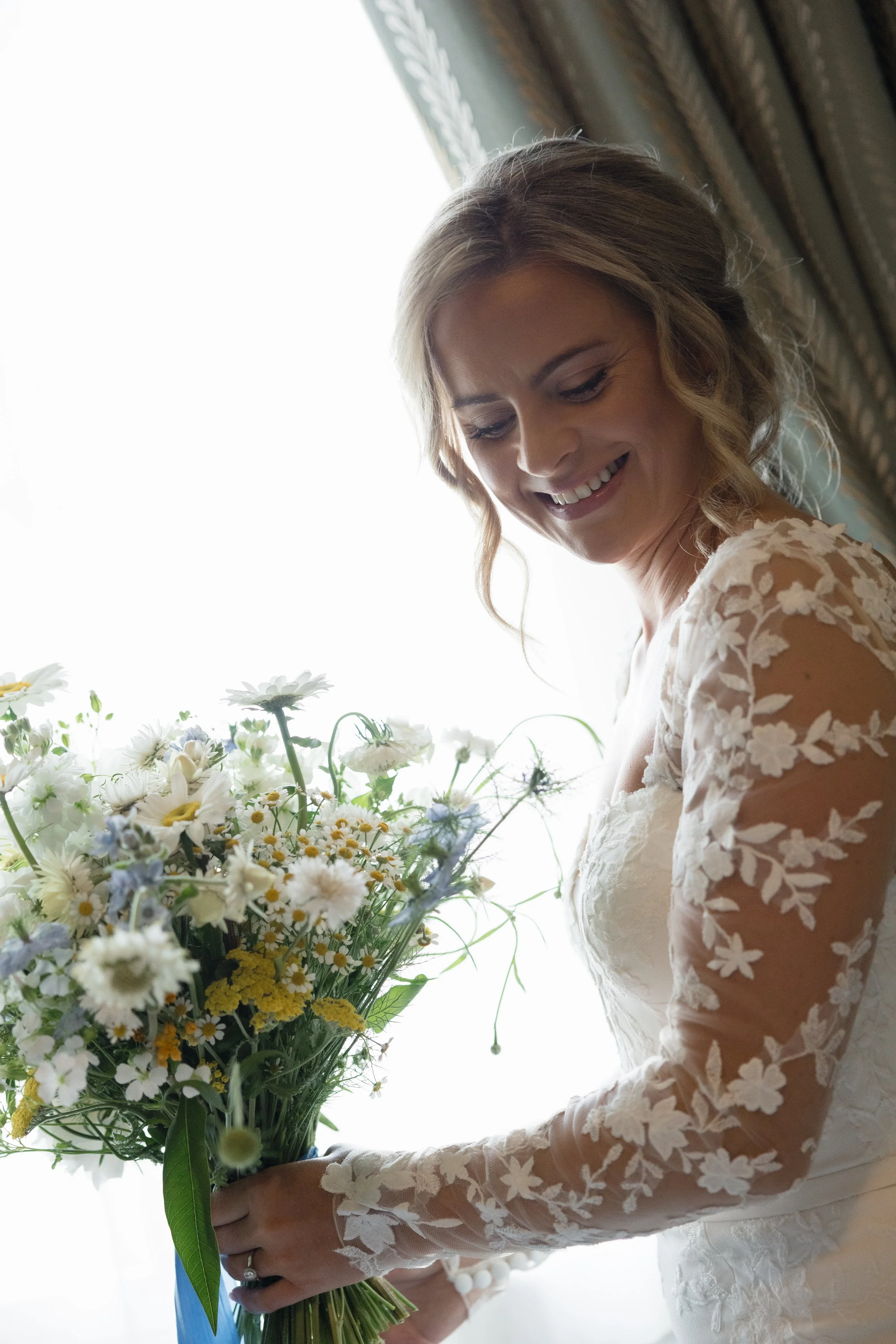 A woman in a wedding dress holding a large bouquet of white, yellow, and purple flowers, smiling while looking down.